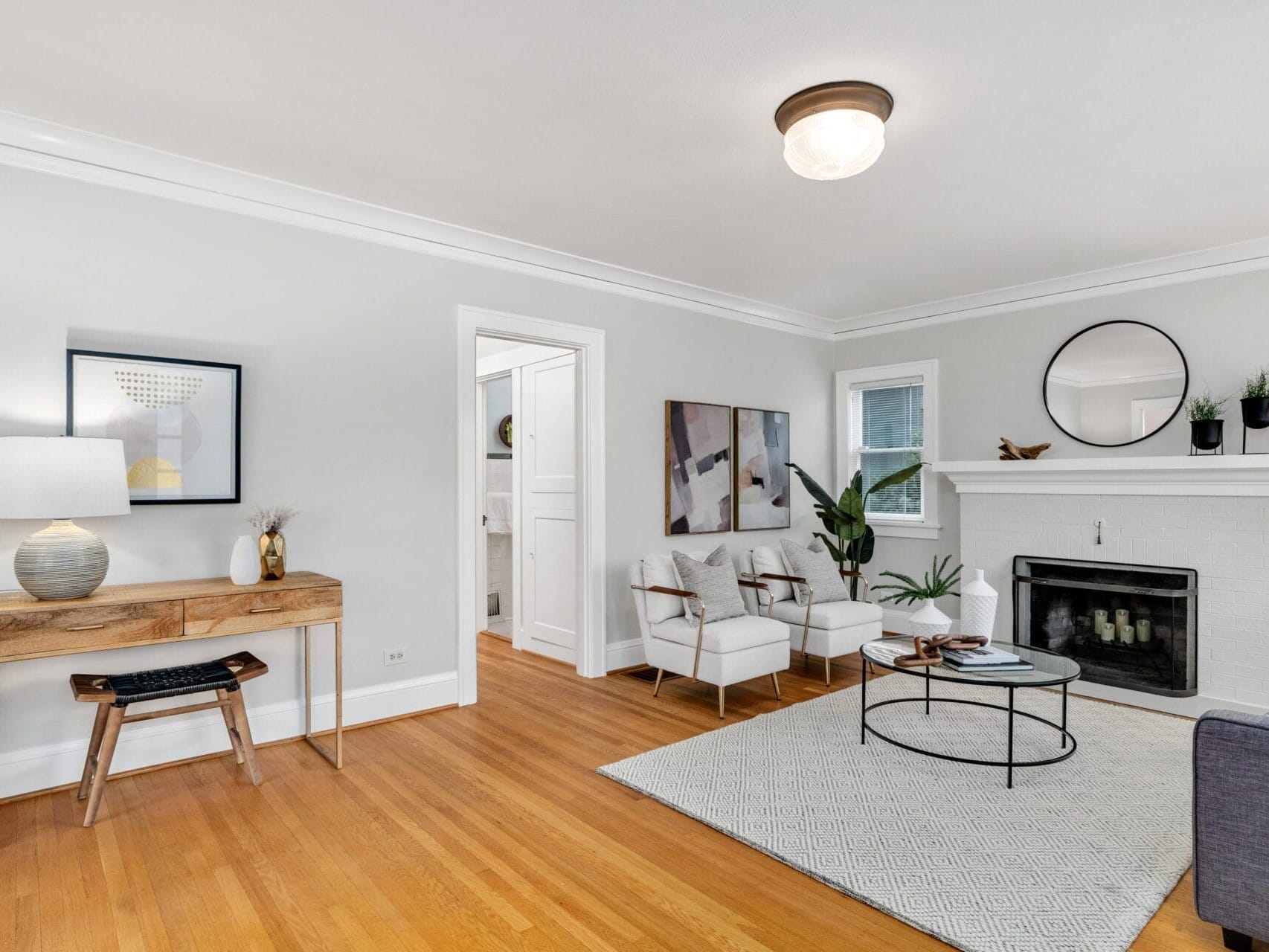 A bright, modern living room featuring a white fireplace, a round mirror, and a cozy sitting area with two white chairs. A wooden desk with a lamp and decor sits on the left, and a large rug covers part of the hardwood floor.