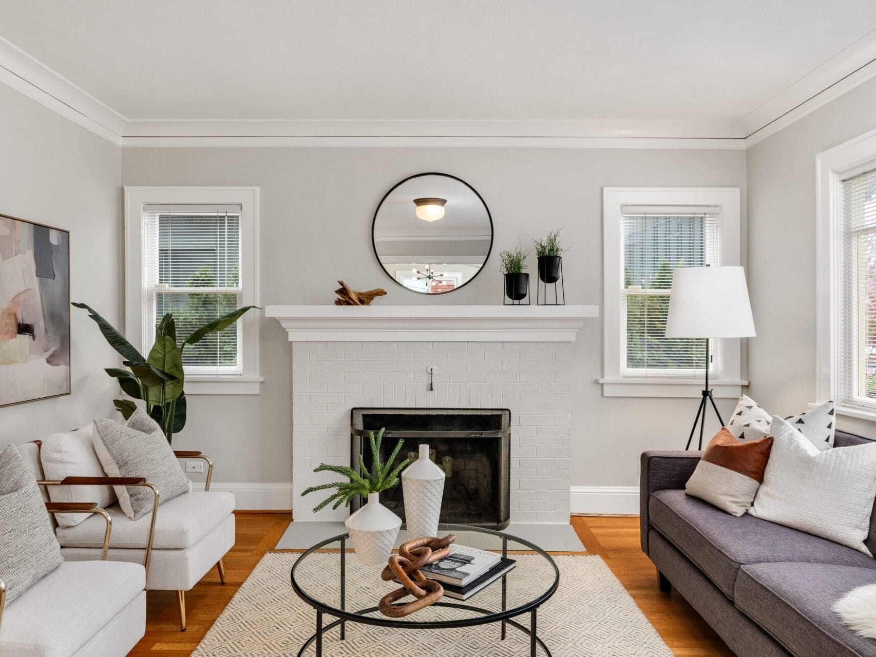 A cozy living room featuring a white brick fireplace with a round mirror above it. There are two white armchairs, a gray sofa with assorted cushions, and a round coffee table. Plants and modern decor add warmth to the space. Natural light fills the room.