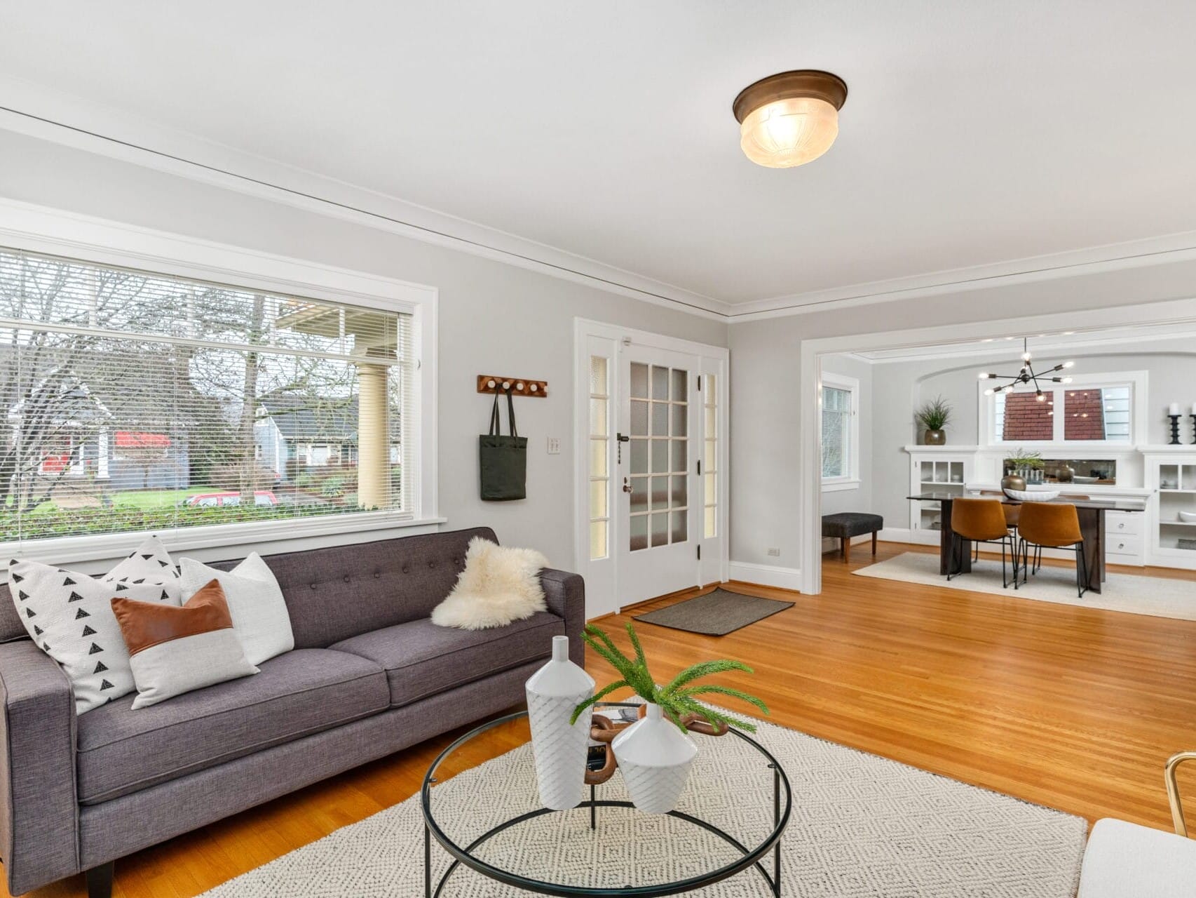 Bright living room with a gray sofa, patterned cushions, a round glass coffee table, and a view into the dining area with white shelves and dark chairs. Large windows let in natural light, and hardwood floors add warmth to the space.