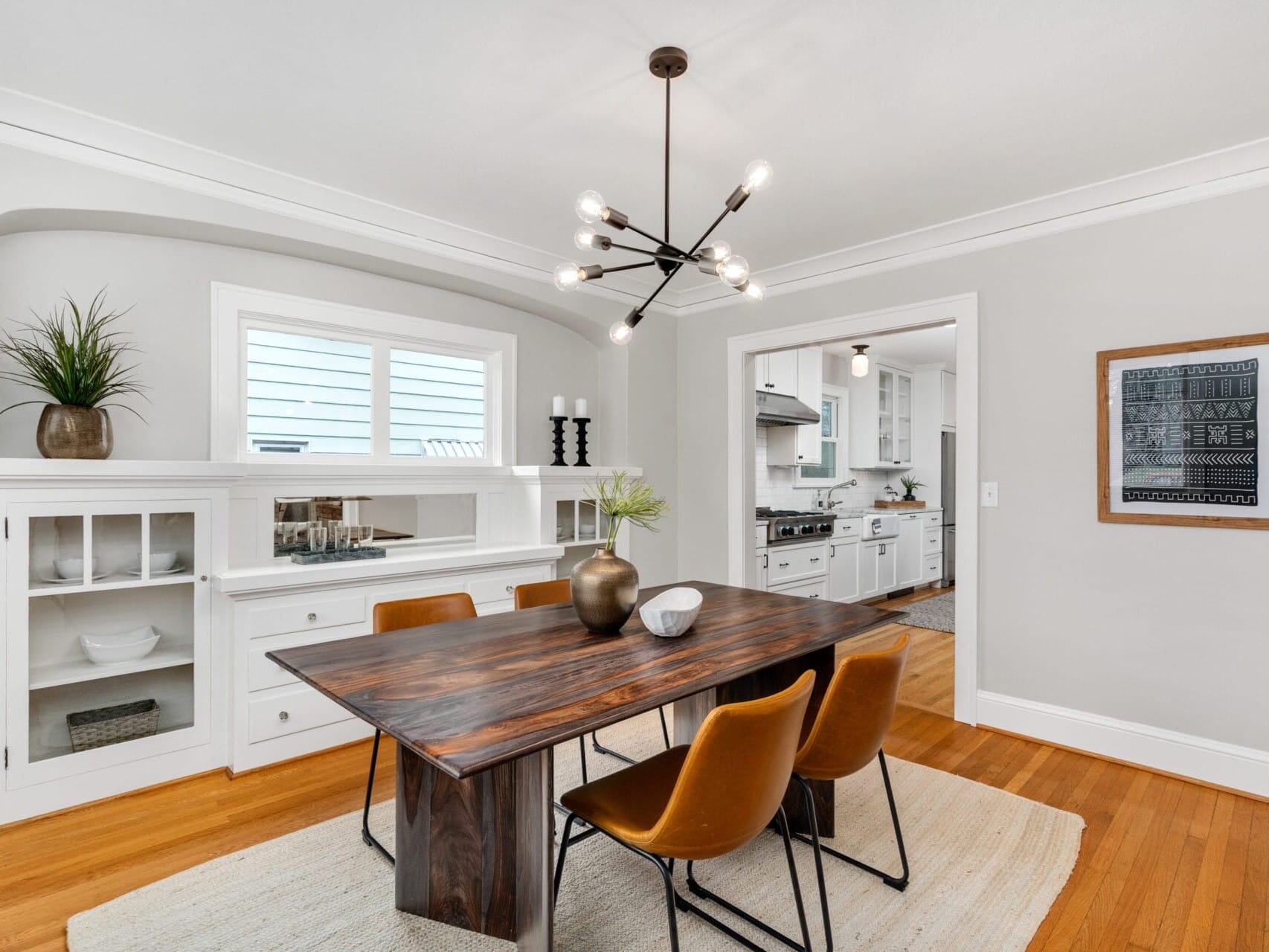 A modern dining room featuring a wooden table with four brown chairs. A geometric chandelier hangs above. Built-in shelves with decor items are on the left. A doorway leads to a white kitchen in the background. Warm wooden floors and a neutral rug complete the space.