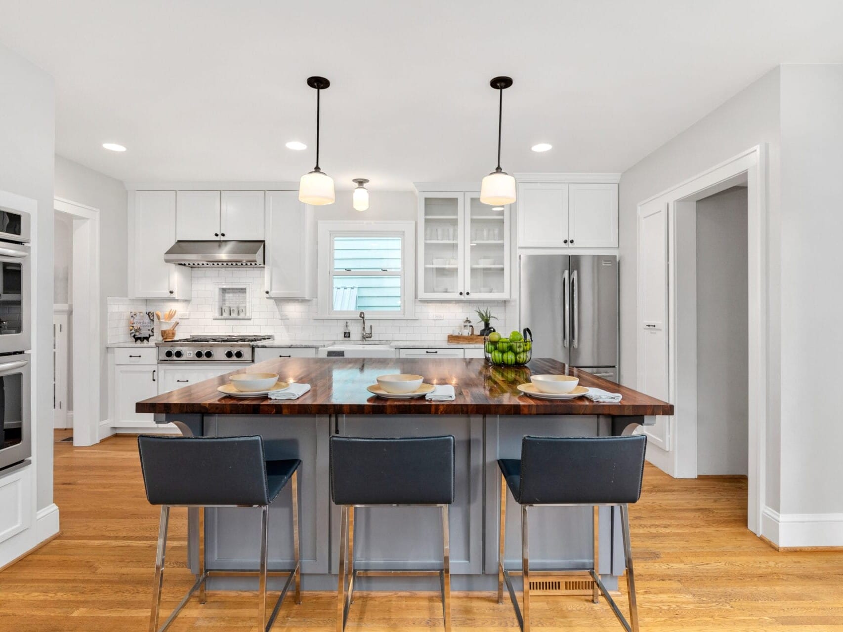 A modern kitchen with white cabinets, stainless steel appliances, and a dark wood island. Three blue chairs are at the island, which is set with plates and bowls. Pendant lights hang above, and hardwood floors complete the look.