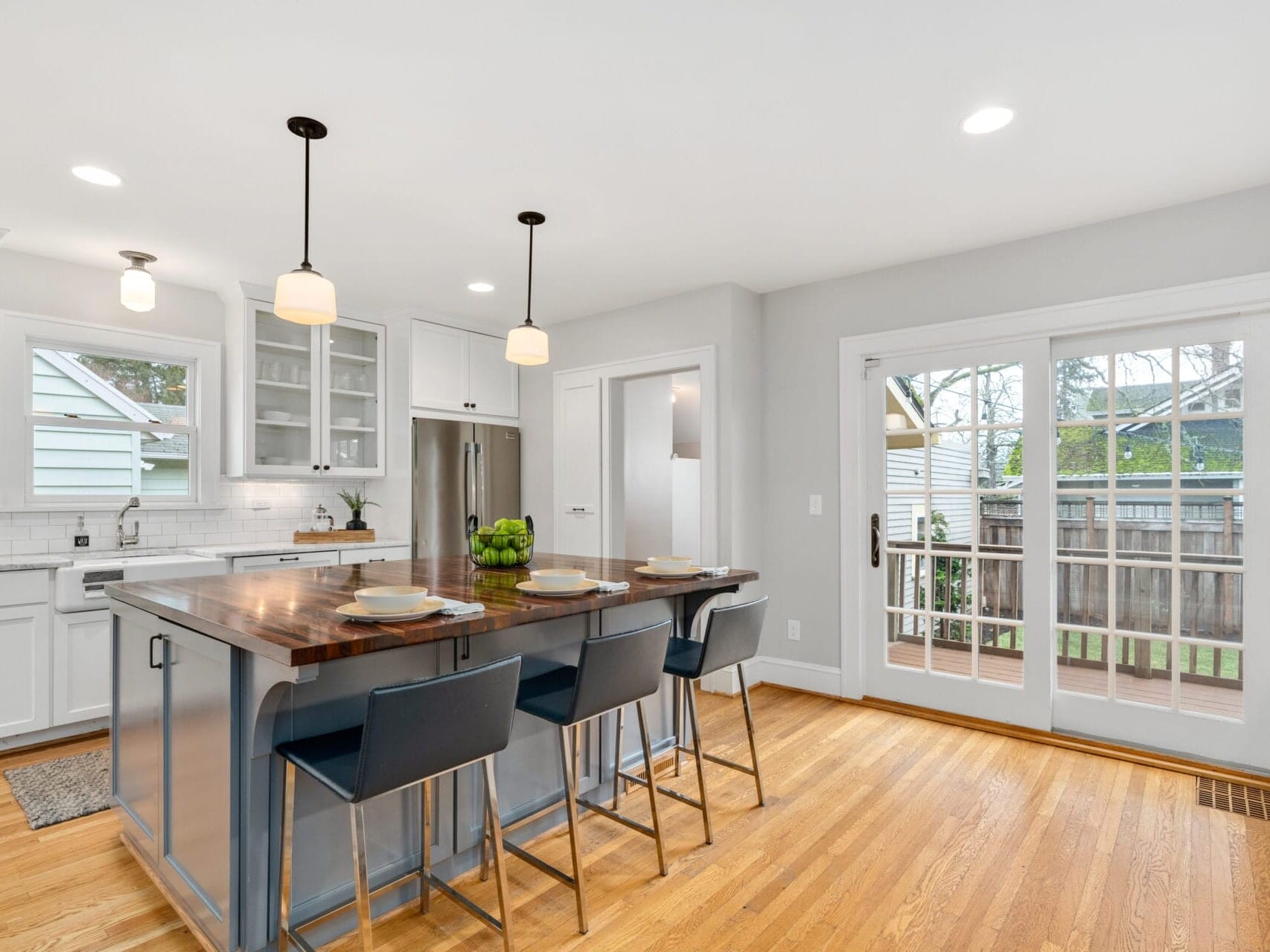 Modern kitchen with white cabinets, hardwood floor, and a central island featuring a dark wood countertop with bar stools. Pendant lights hang above. Sliding glass doors open to a deck with a view of a fence and trees outside.