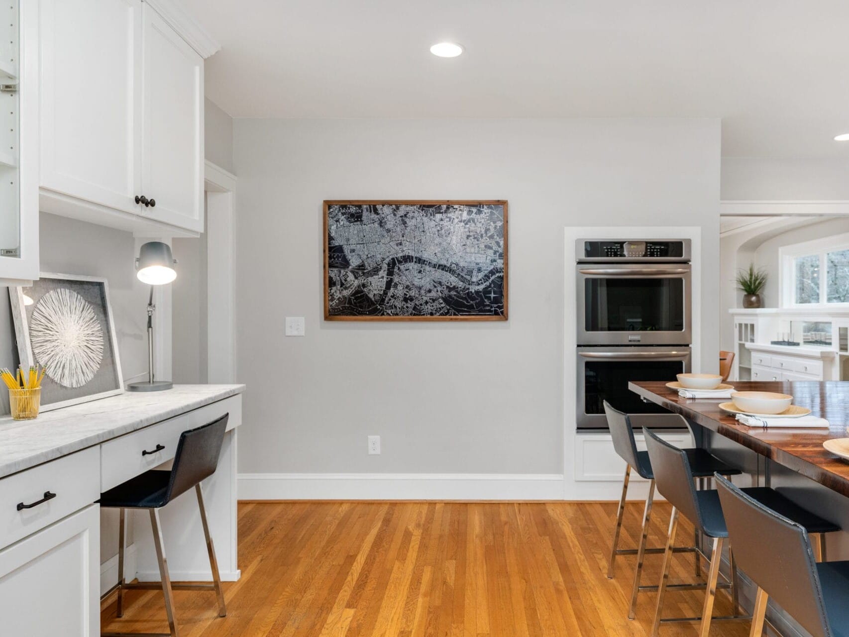 A modern kitchen with wooden floors, white cabinets, and black chairs. A marble countertop has a desk lamp and a pencil holder. The dining table is set with plates and bowls. A map artwork hangs on the wall above a double oven. Natural light fills the room.