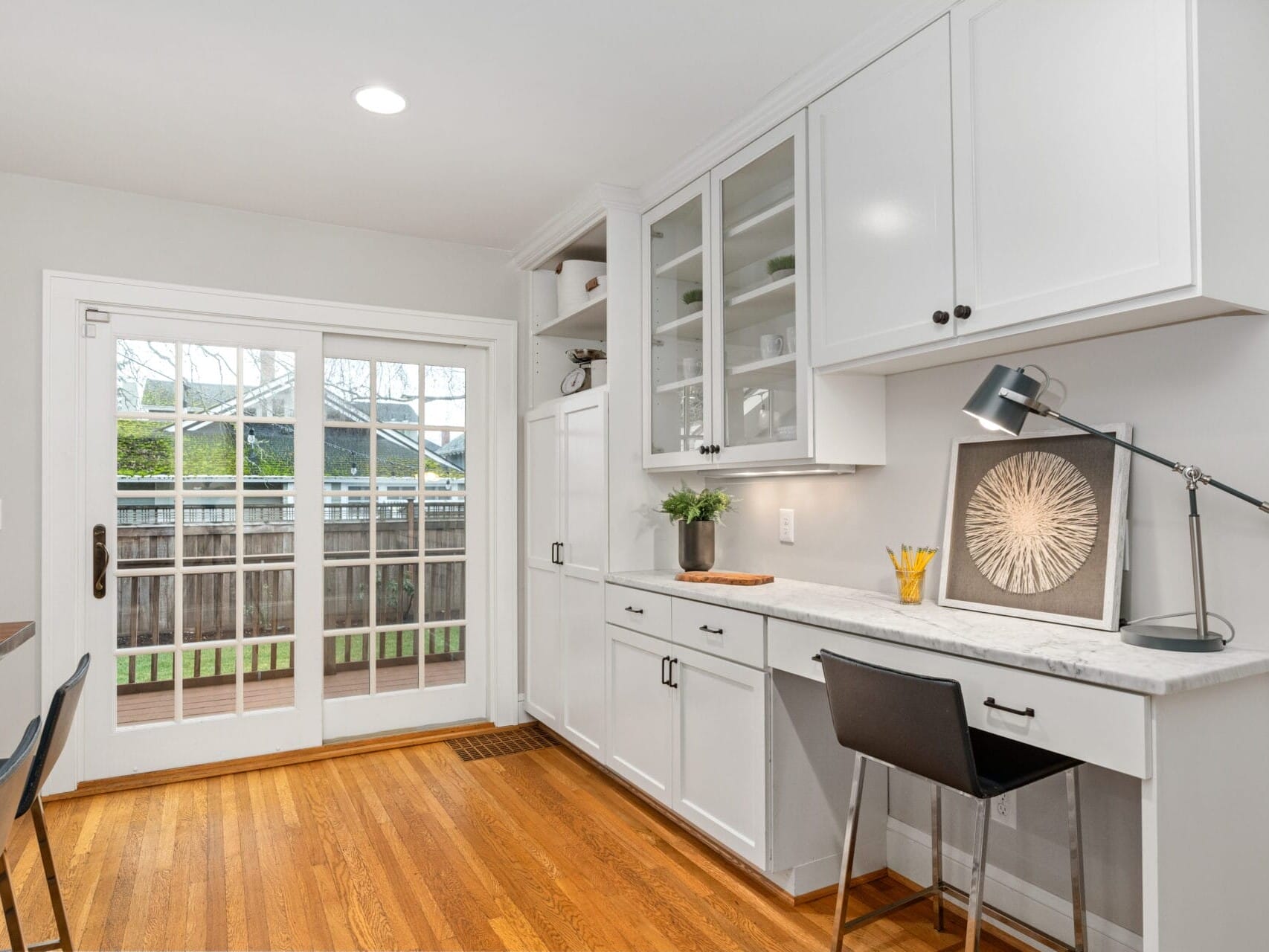 A home office space in a kitchen features white cabinets, a built-in desk with a chair, and decorative items. The room has wooden floors and French doors leading to a patio, creating a bright and functional workspace.