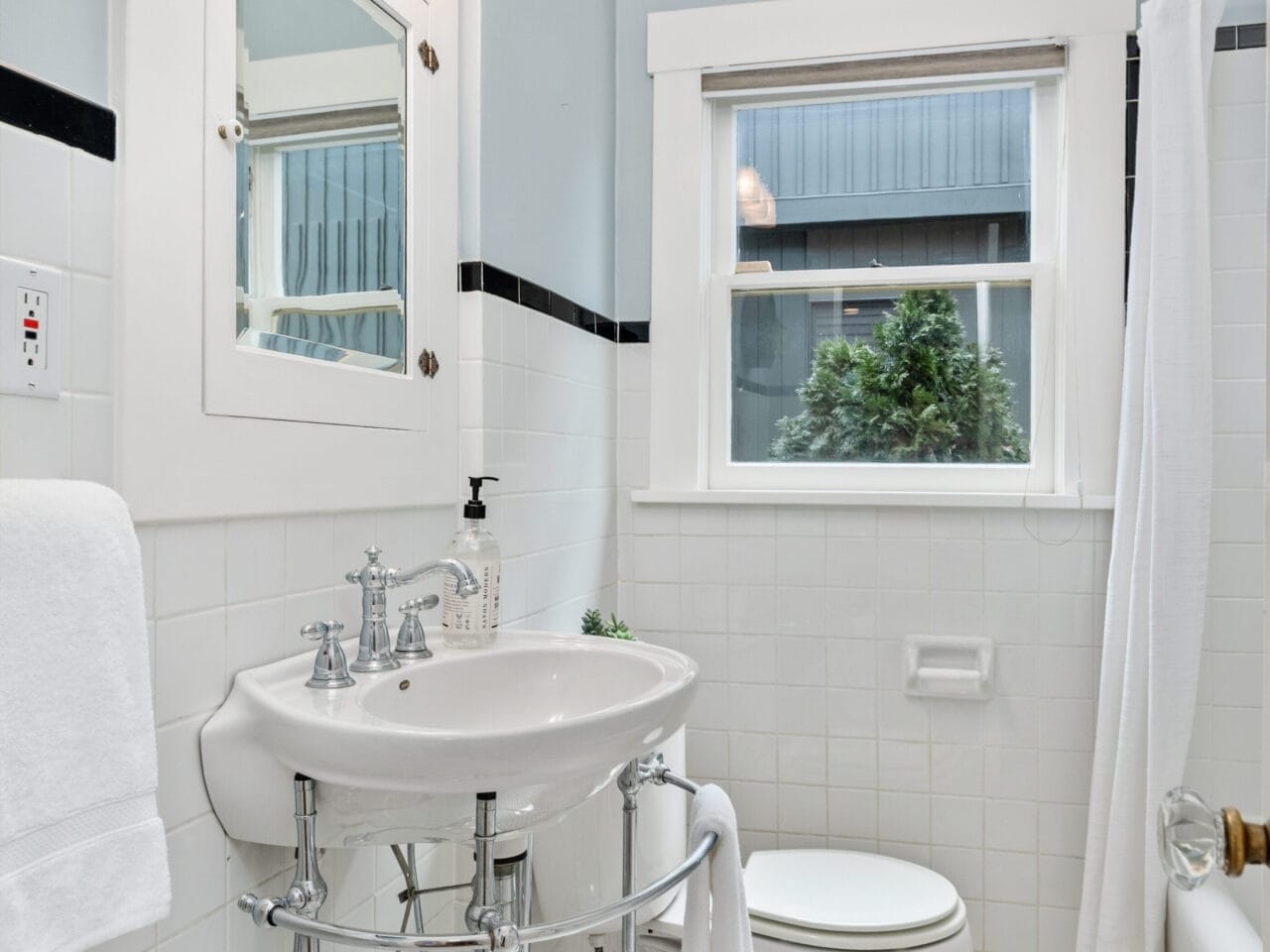 Small, bright bathroom with light blue walls, white tiles, and a hexagonal tile floor. Features a wall-mounted sink with an oval mirror above, a toilet, and a bathtub with a shower curtain. A window allows natural light in.