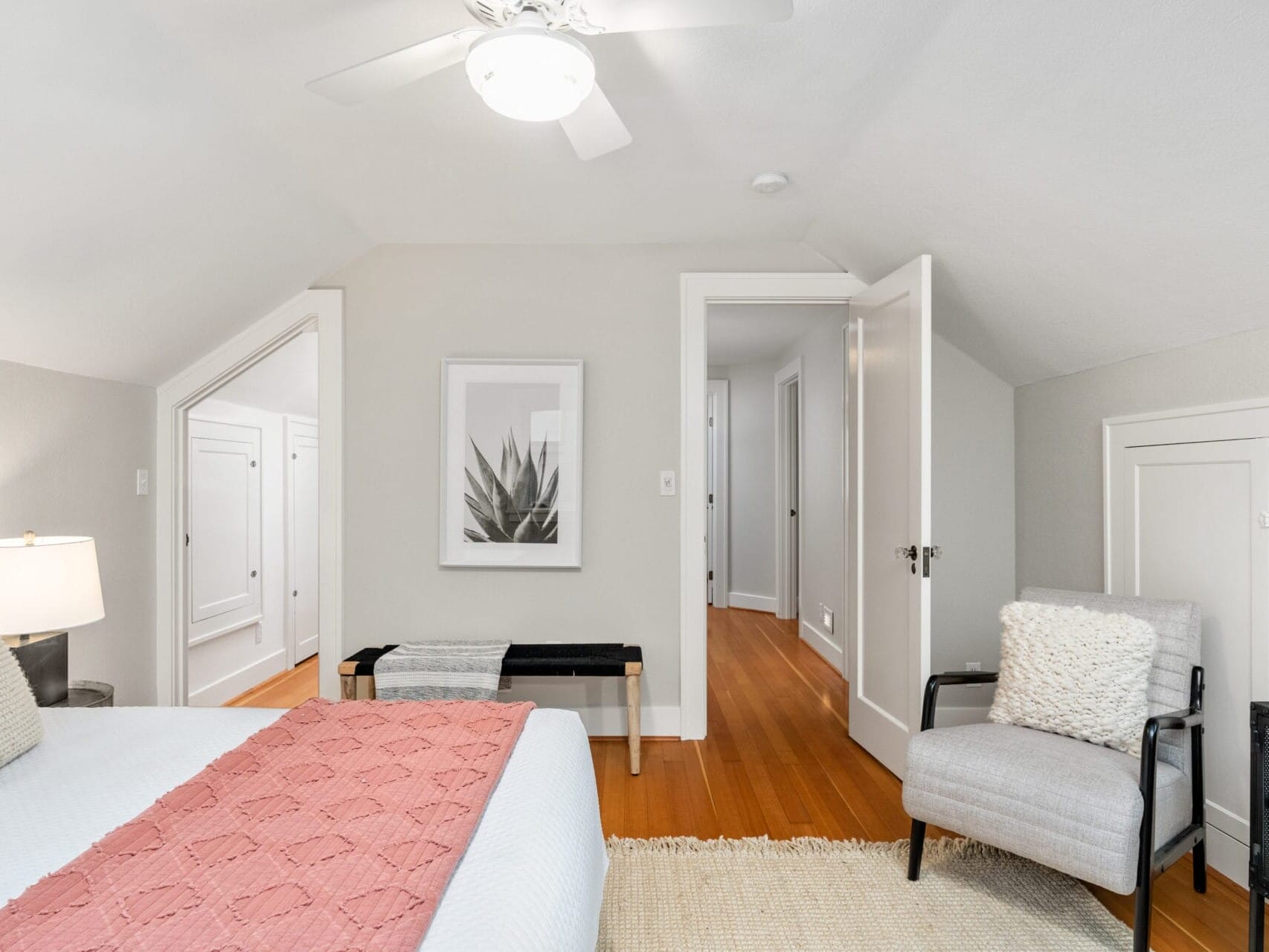 A cozy, modern bedroom with a sloped ceiling. It features a bed with a pink blanket, a gray armchair, a small bench, and a minimalist plant artwork on the wall. The room is well-lit with a ceiling fan light and has hardwood floors.