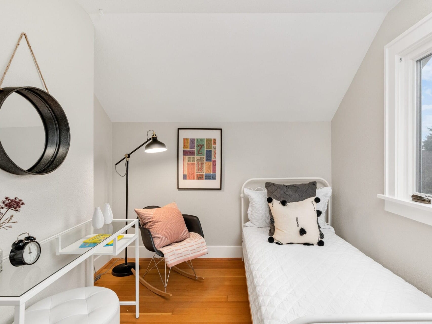 A small, cozy bedroom with a single bed, white bedding, and decorative pillows. A desk with a chair, lamp, clock, and plants sits near the bed. A round mirror hangs on the wall, and a colorful poster adds a touch of vibrancy. A window provides natural light.