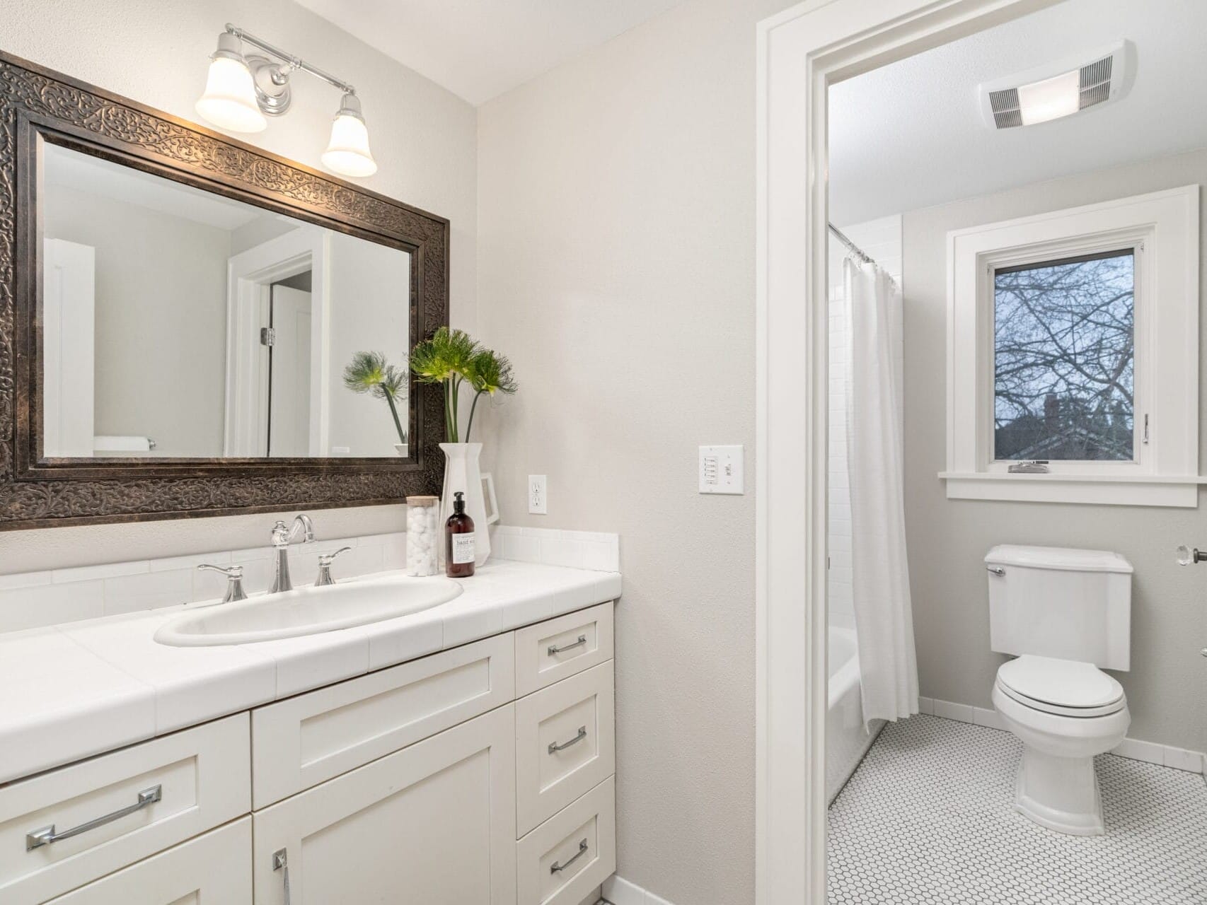 A bright, modern bathroom features a white vanity with a large framed mirror, a sink with dual faucets, and a potted plant. A toilet is next to a window with a view of tree branches, and the floor is covered with small patterned tiles.