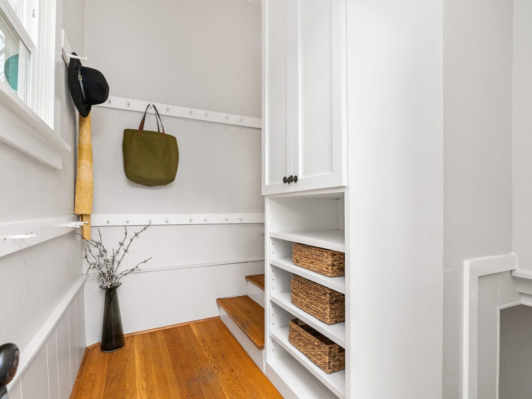 A small mudroom with wooden flooring, featuring white walls and shelves. A hat, green bag, and yellow scarf hang on wall hooks. Baskets are stored in open shelves. A vase with branches decorates the corner near a window.