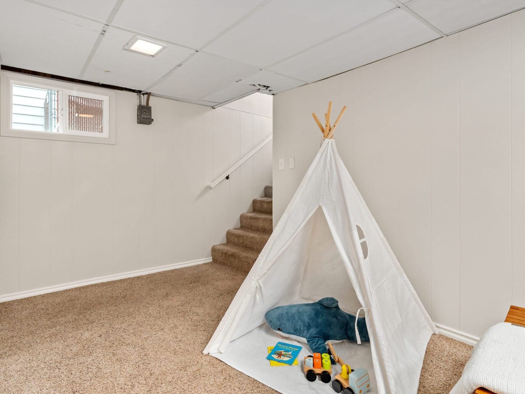 A carpeted basement room with white walls features a childrens play area. A small white teepee tent with toys—an inflatable dolphin, a stuffed animal, and a toy truck—is set up. A staircase leads to the upper level, and a single window allows natural light in.