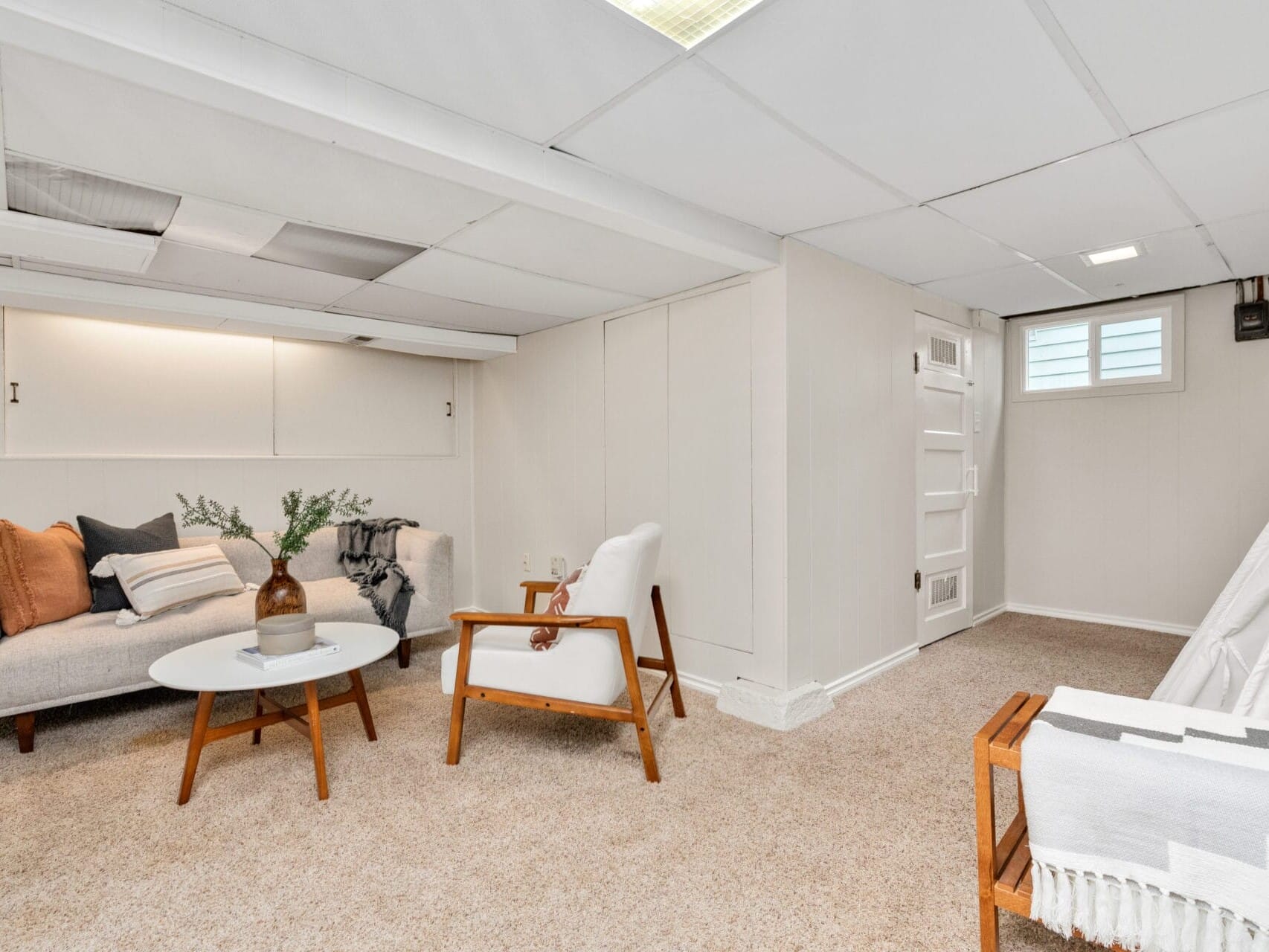 A cozy basement living area with beige carpet, featuring a gray couch with cushions, a round white coffee table, two armchairs, and a small window. A decorative vase with greenery sits on the table, and a blanket is draped over an armchair.