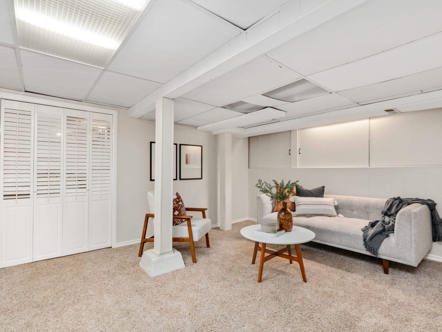 A minimalist living room with a gray couch, two wooden armchairs, and a round white coffee table. The walls are light-colored, and the ceiling has recessed lighting. A small potted plant and decorative items are on the table. Carpeted floor.