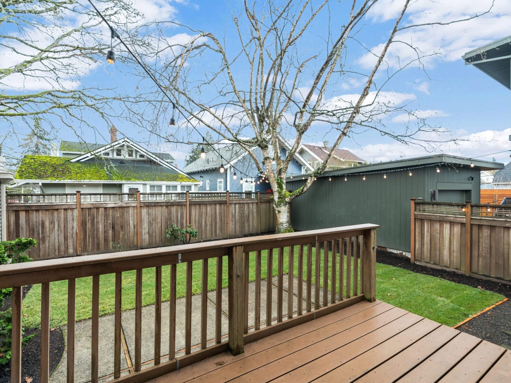 A wooden deck overlooks a small fenced backyard with a large bare tree. The yard features a grassy area and a paved path. Neighboring houses with blue and gray exteriors are visible in the background under a clear blue sky.