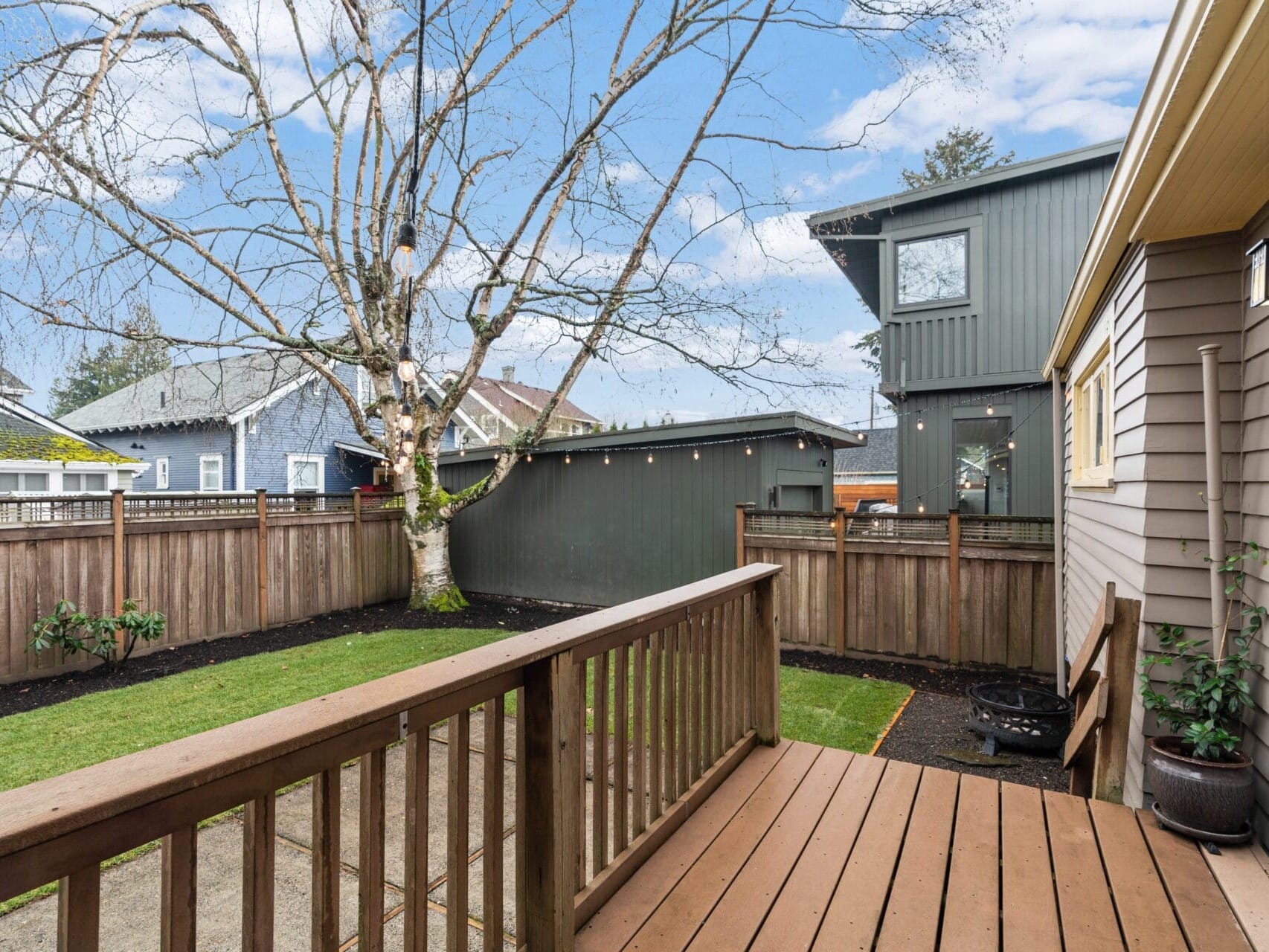 A wooden deck overlooking a small backyard with a tree and grass. Fenced area with neighboring houses visible. The sky is blue with a few clouds. Cozy outdoor space with string lights along the fence.