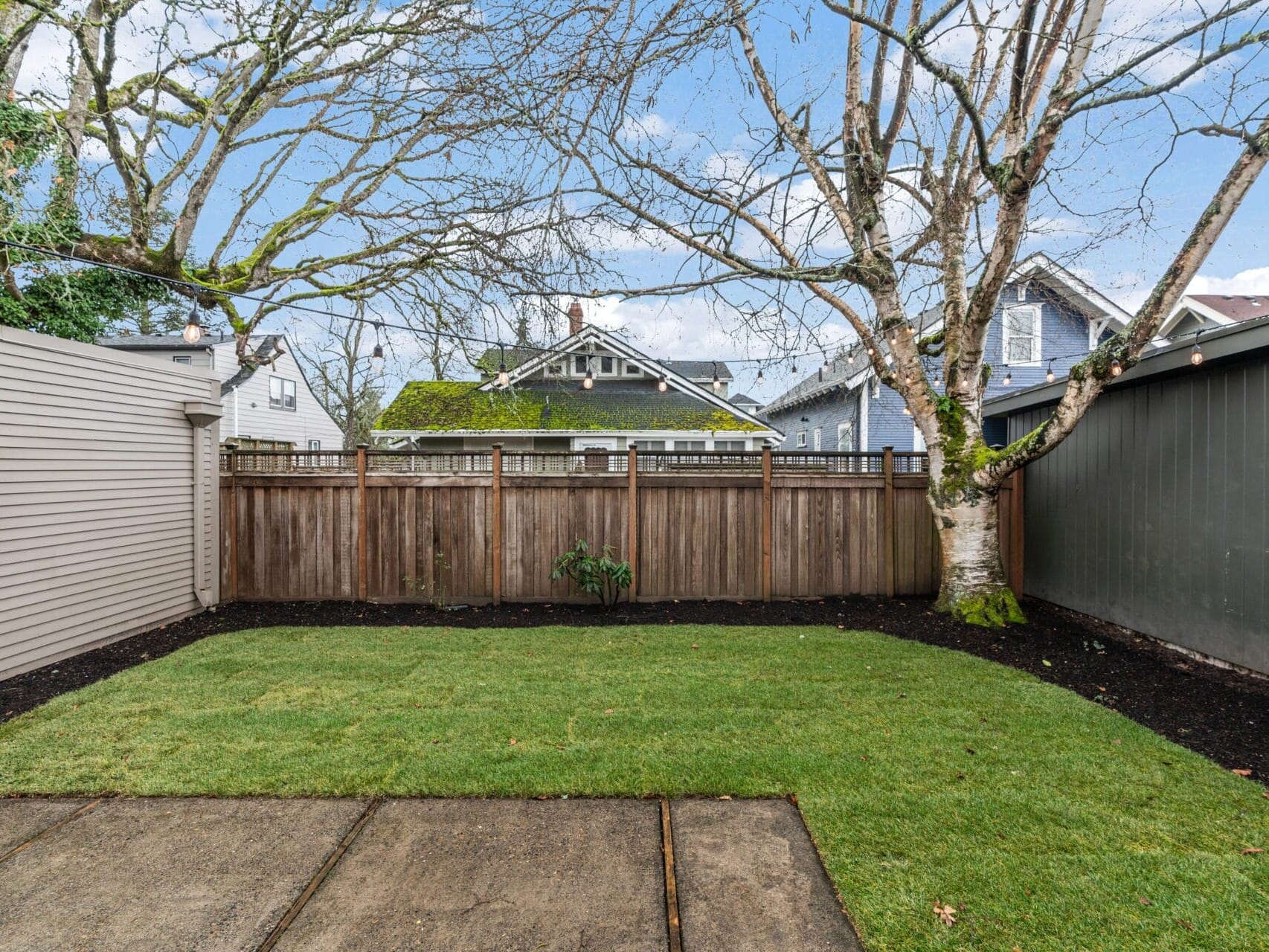 A backyard with a small, grassy lawn surrounded by wooden fencing. Trees with bare branches overhang, and there are two small shrubs along the fence. The sky is partly cloudy, and the surrounding houses are visible in the background.