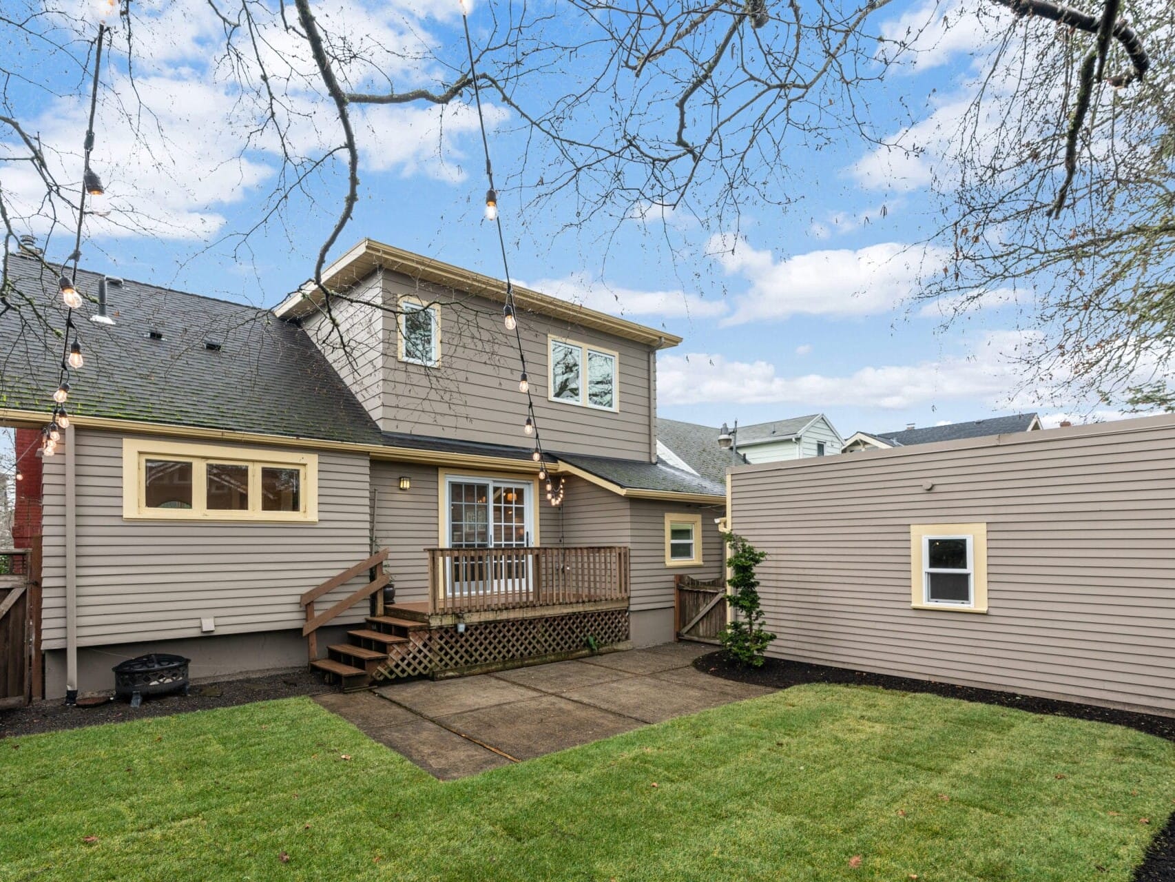 A backyard view of a two-story house with taupe siding and white trim. It features a small wooden deck with stairs leading to a neatly trimmed lawn. String lights hang above, and a tall fence surrounds the yard. The sky is partly cloudy.