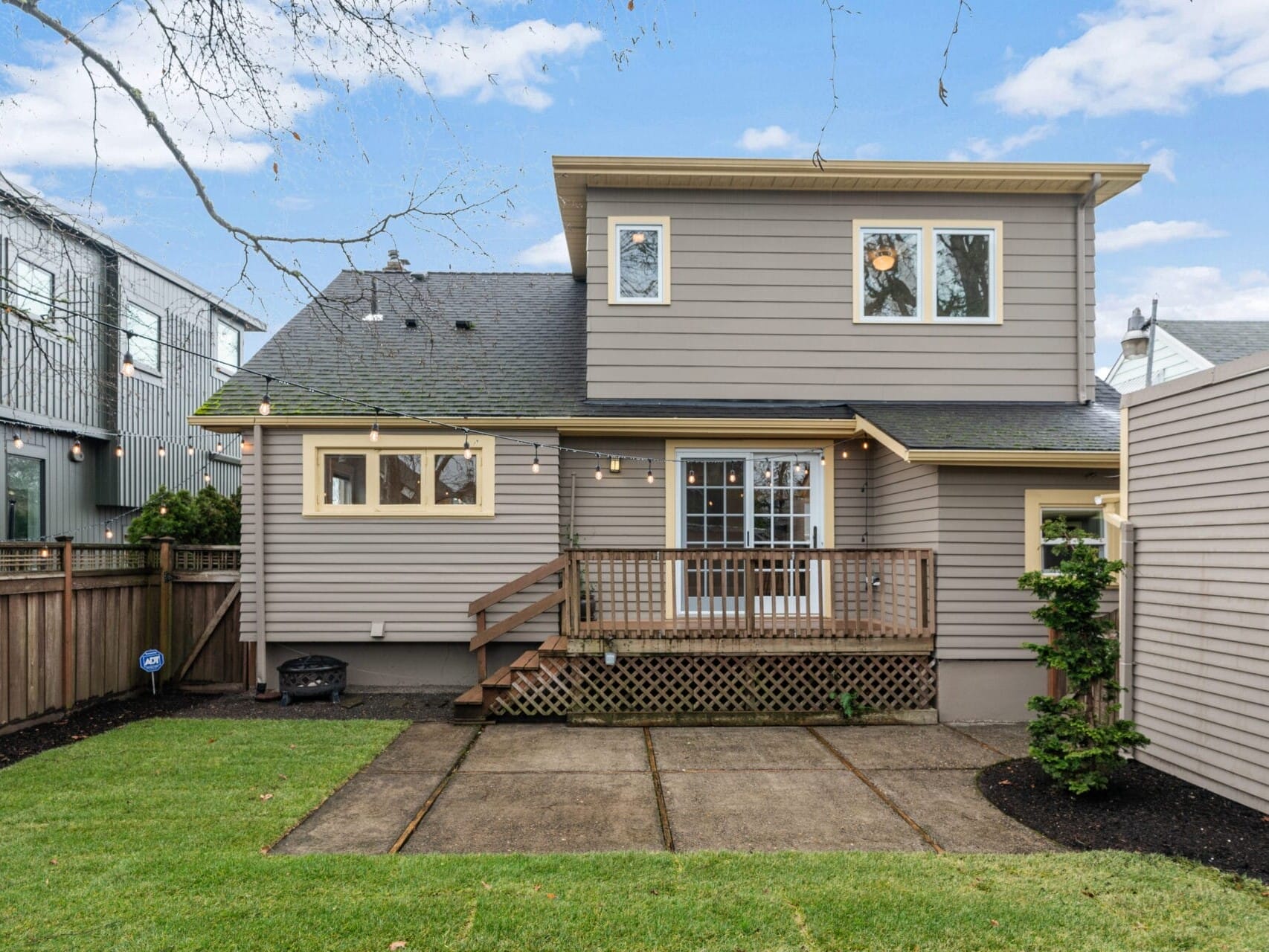 The image shows the back of a two-story house with gray siding and a small wooden deck with stairs leading down to a concrete patio. The yard has a neatly trimmed lawn, surrounded by a wooden fence. Outdoor string lights are hung above the deck.