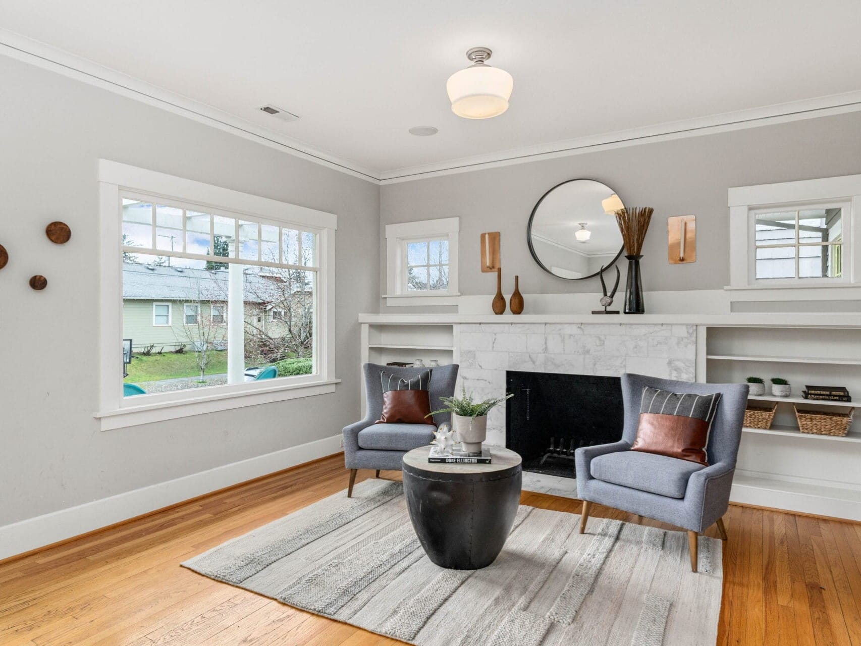 A cozy living room with two gray armchairs facing a round table on a rug. A white fireplace is centered between built-in shelves, holding decorative items. A round mirror hangs above the mantel, and large windows let in natural light.