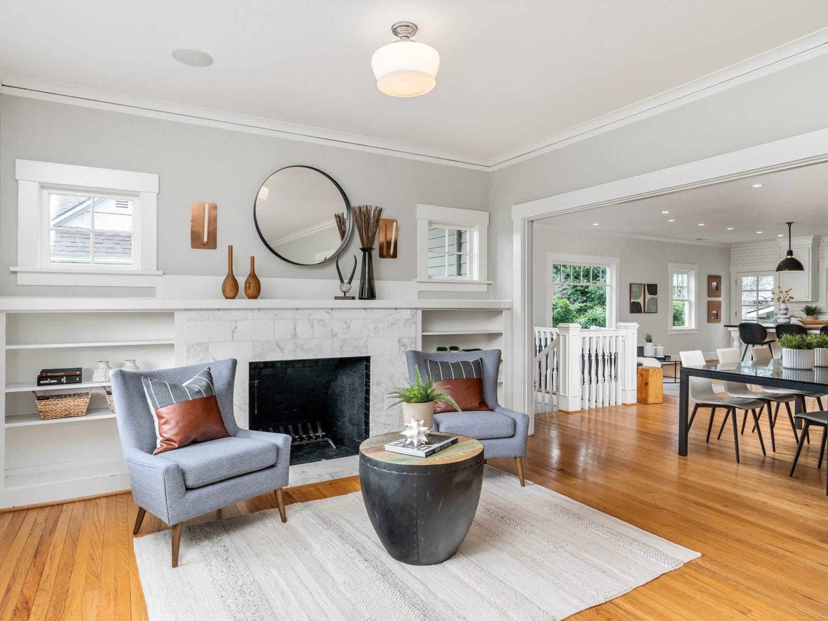 A modern living room with hardwood floors features two gray chairs and a round black coffee table on a light rug. A fireplace is adorned with a round mirror and decorative items. The dining area is visible in the background. Natural light fills the space.