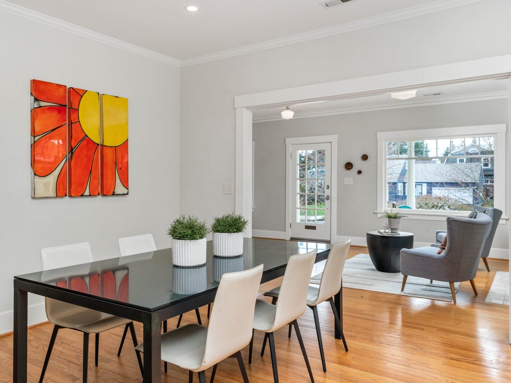 A modern dining room with a black table and six white chairs on a wooden floor. A large, colorful flower painting is on the wall. In the background, a living area with a gray armchair, small table, and potted plant is visible.