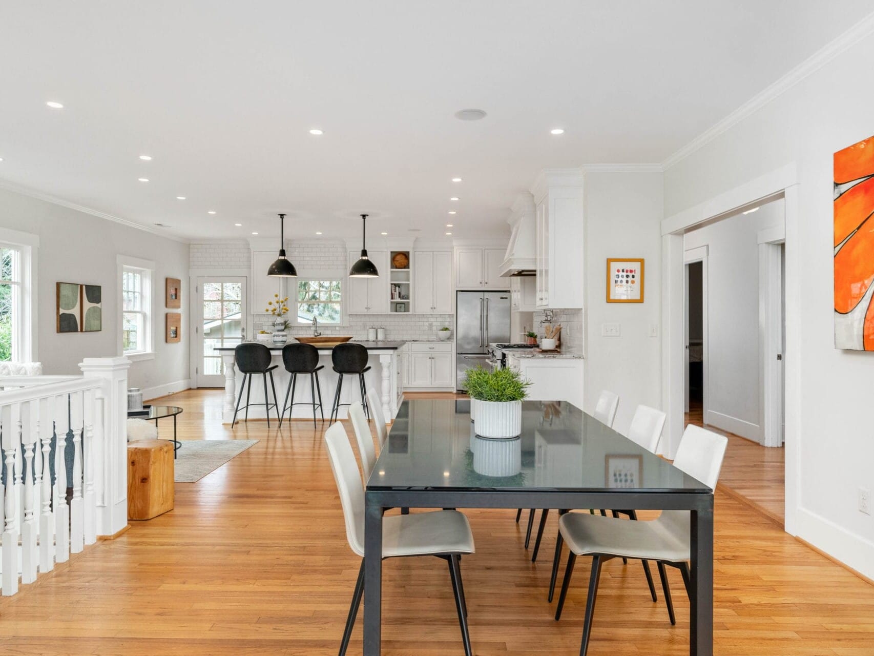 A modern, open-concept kitchen and dining area with wooden floors. The dining table has six white chairs and a potted plant centerpiece. The kitchen features bar stools, white cabinets, and stainless steel appliances. Bright artwork hangs on the wall.