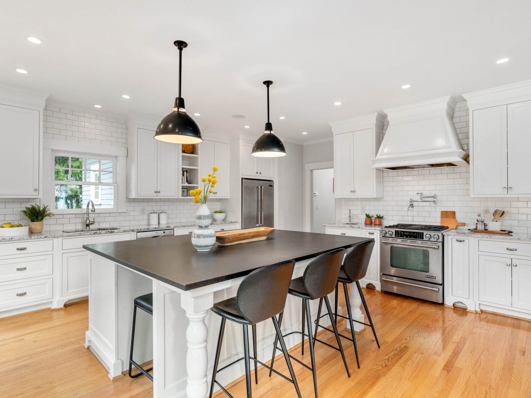 Bright, modern kitchen with white cabinets, a large central island with black countertops, and three black barstools. Stainless steel appliances and pendant lights above the island. Wood flooring adds warmth to the space.