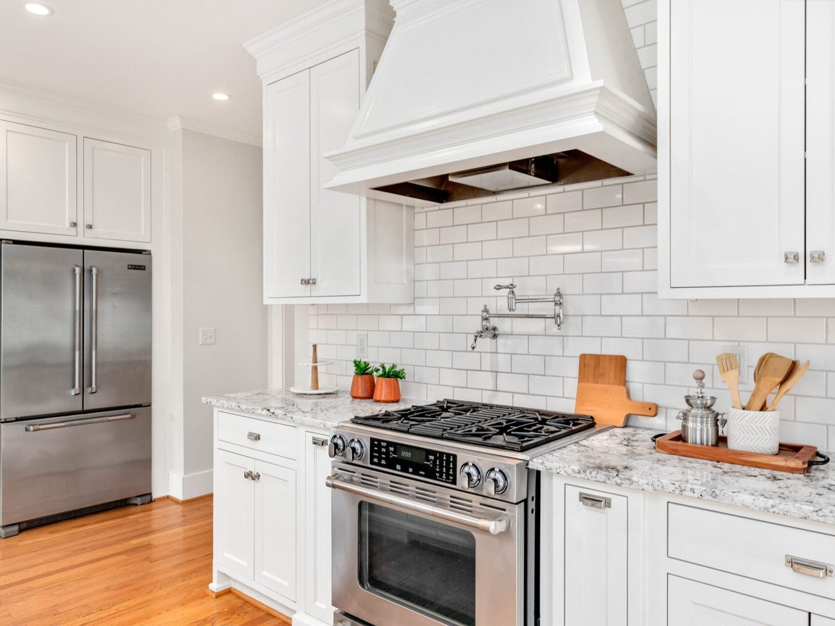 A modern kitchen with white cabinets and a marble countertop. A stainless steel oven is centered beneath a white range hood. A fridge is visible to the left. Wooden utensils and a cutting board are placed on the counter, with potted plants for decor.