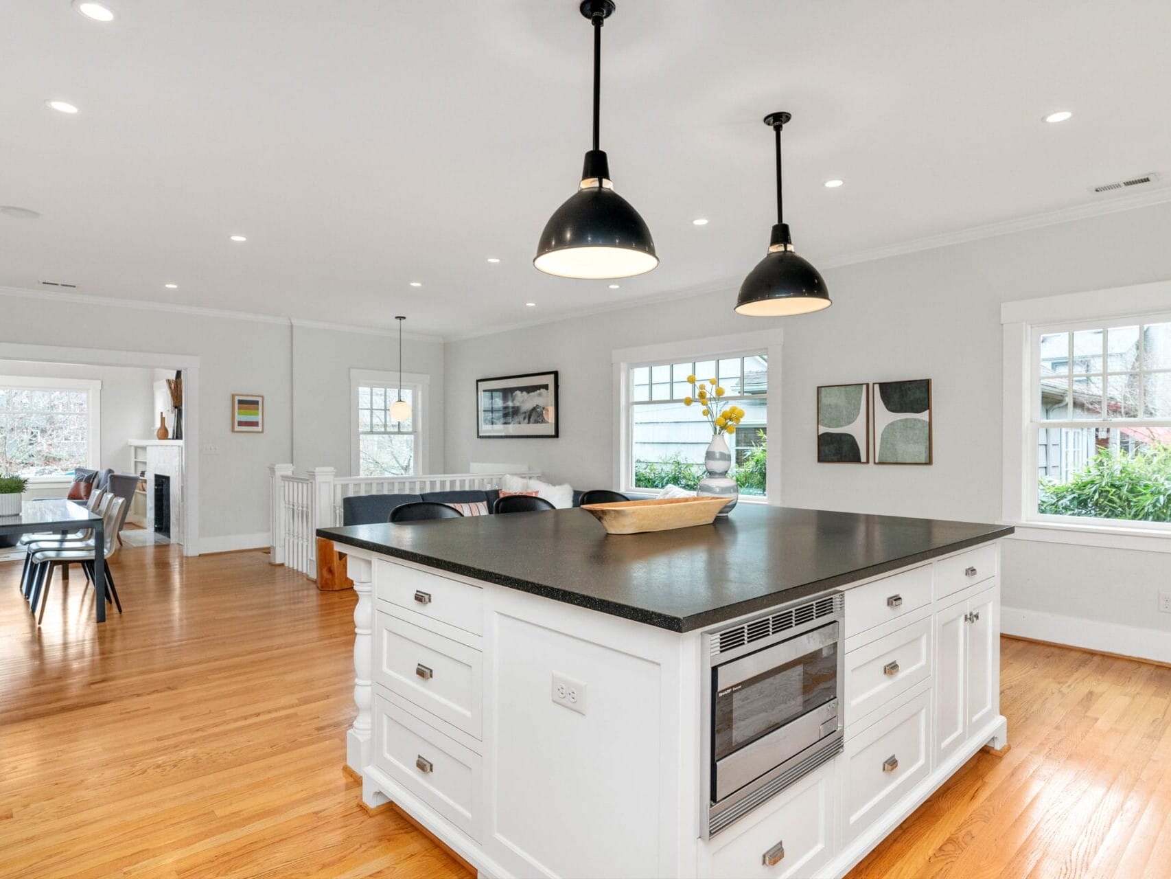 A spacious modern kitchen with a large black countertop island on a wooden floor. Two black pendant lights hang above. In the background, theres a dining table and large windows letting in natural light. White walls and various framed artworks are visible.