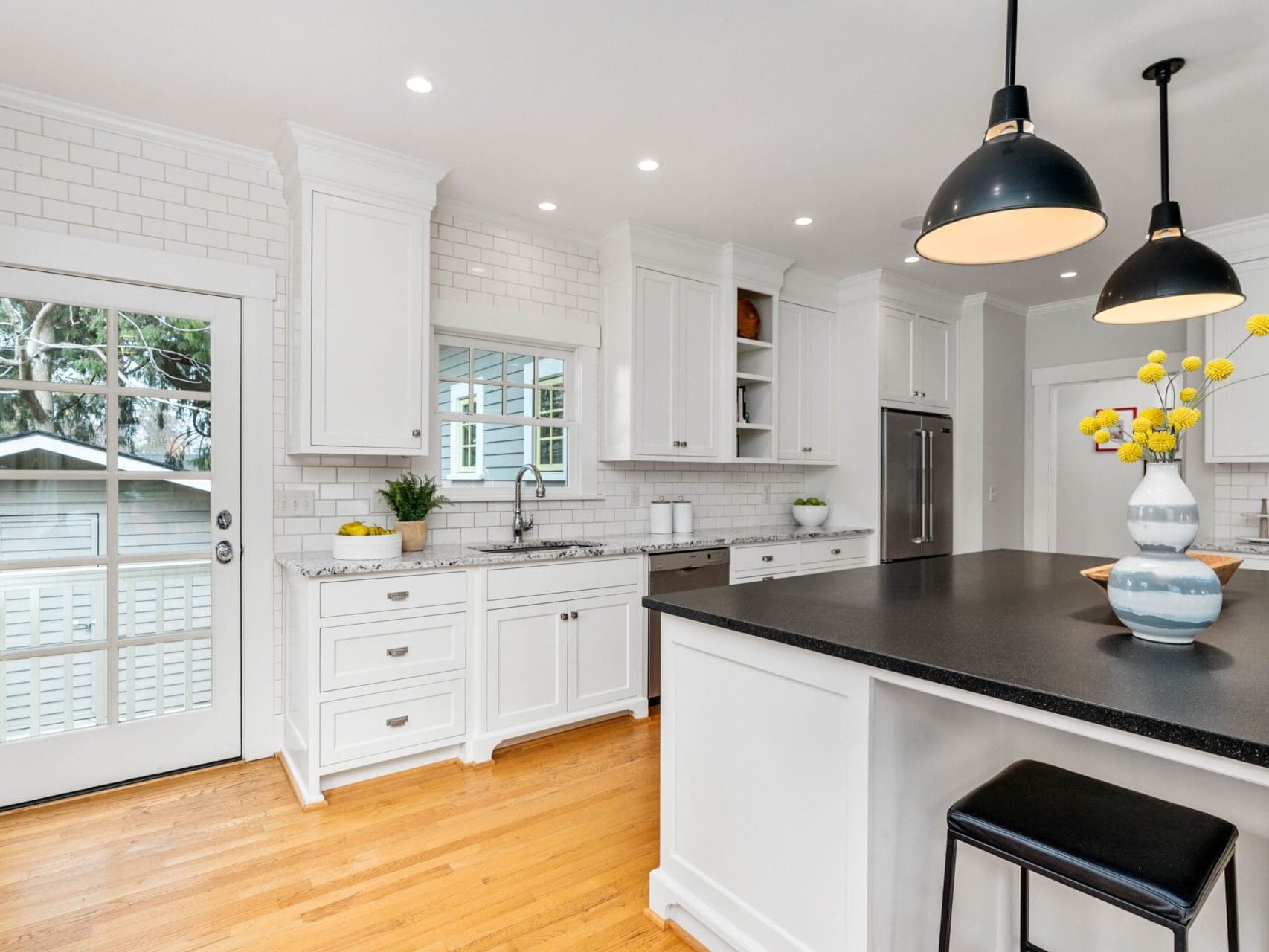 A bright, modern kitchen with white cabinets and a black countertop island. Two pendant lights hang over the island, which has a vase of yellow flowers. A windowed door on the left leads outside. The floor is wooden, and the backsplash is white subway tile.