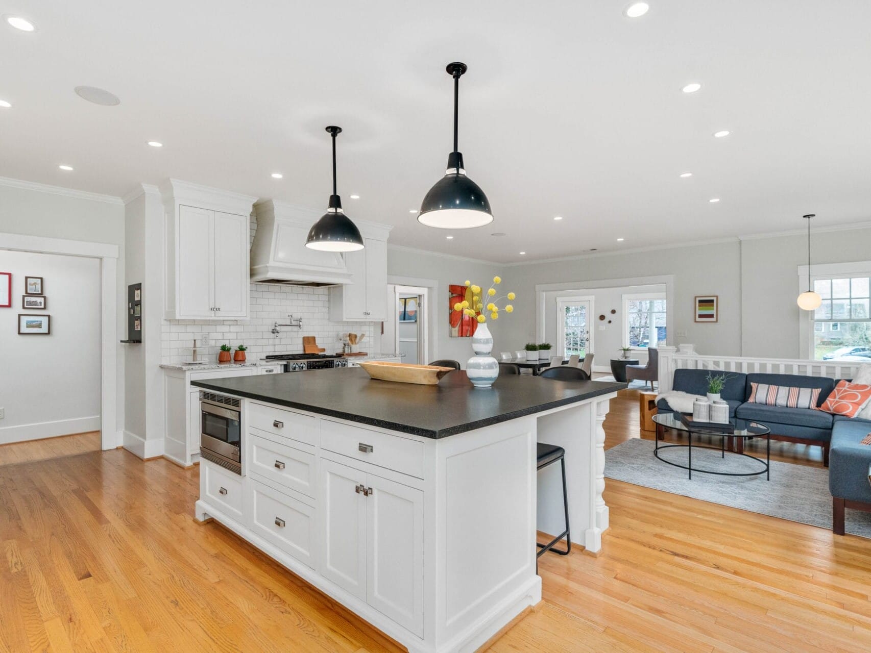 Spacious modern kitchen with hardwood floors, white cabinetry, black countertops, and two large pendant lights. An island with seating divides the kitchen from the living area, which features a sofa, coffee table, and various decorations. Natural light fills the space.