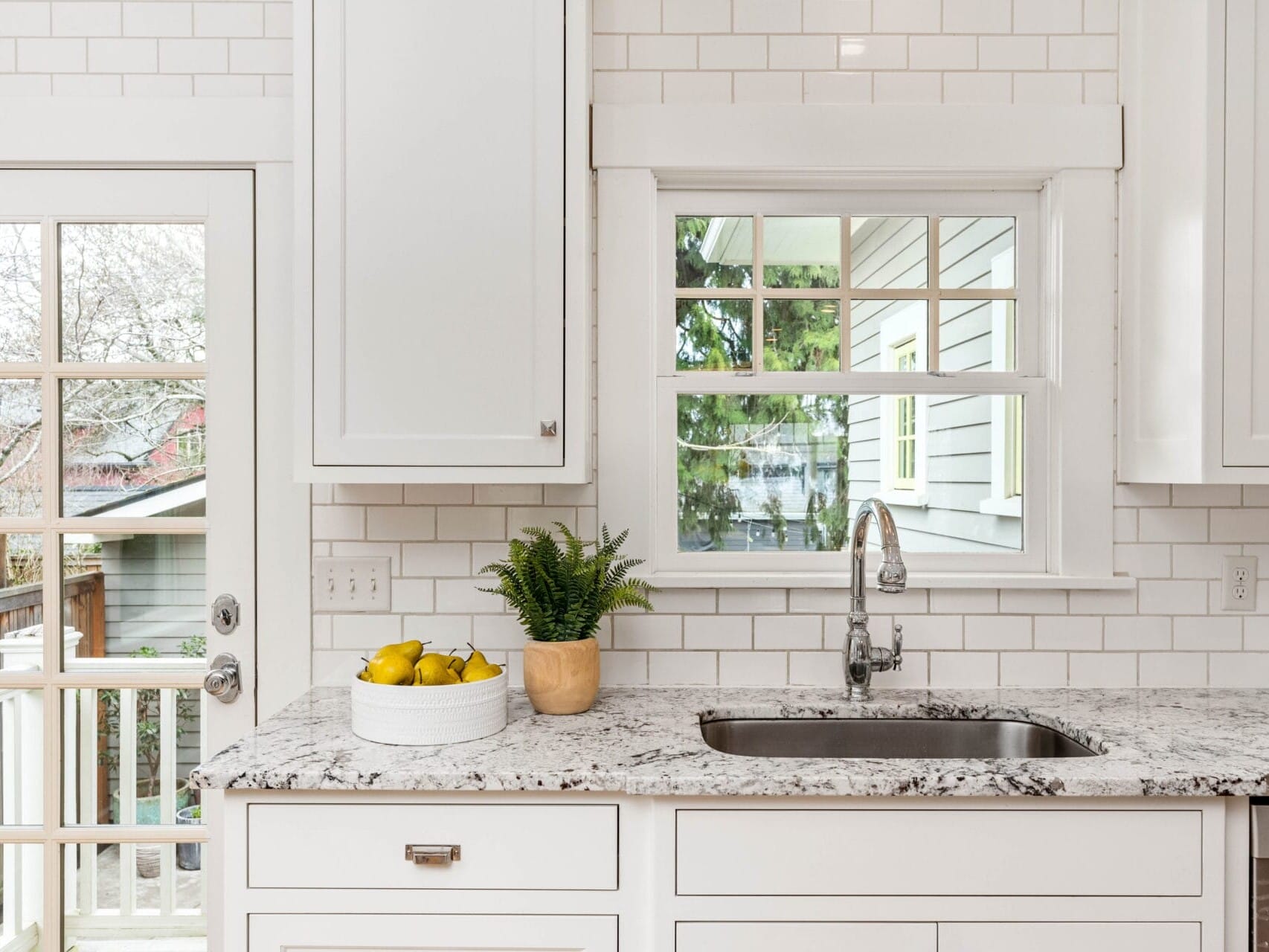 A bright kitchen with white cabinets, a marble countertop, and a stainless steel sink. Theres a window above the sink, a potted plant, and a bowl of lemons. A glass door opens to a patio.