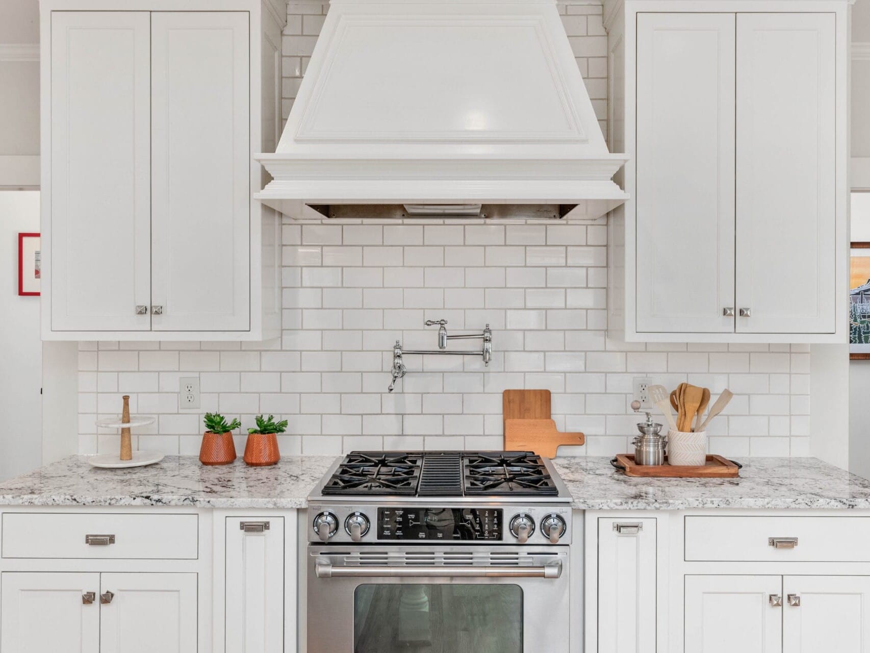 A bright kitchen with white cabinets and a subway tile backsplash. A stainless steel oven is centered below a large white range hood. Marble countertops display potted plants, a cutting board, and kitchen utensils.