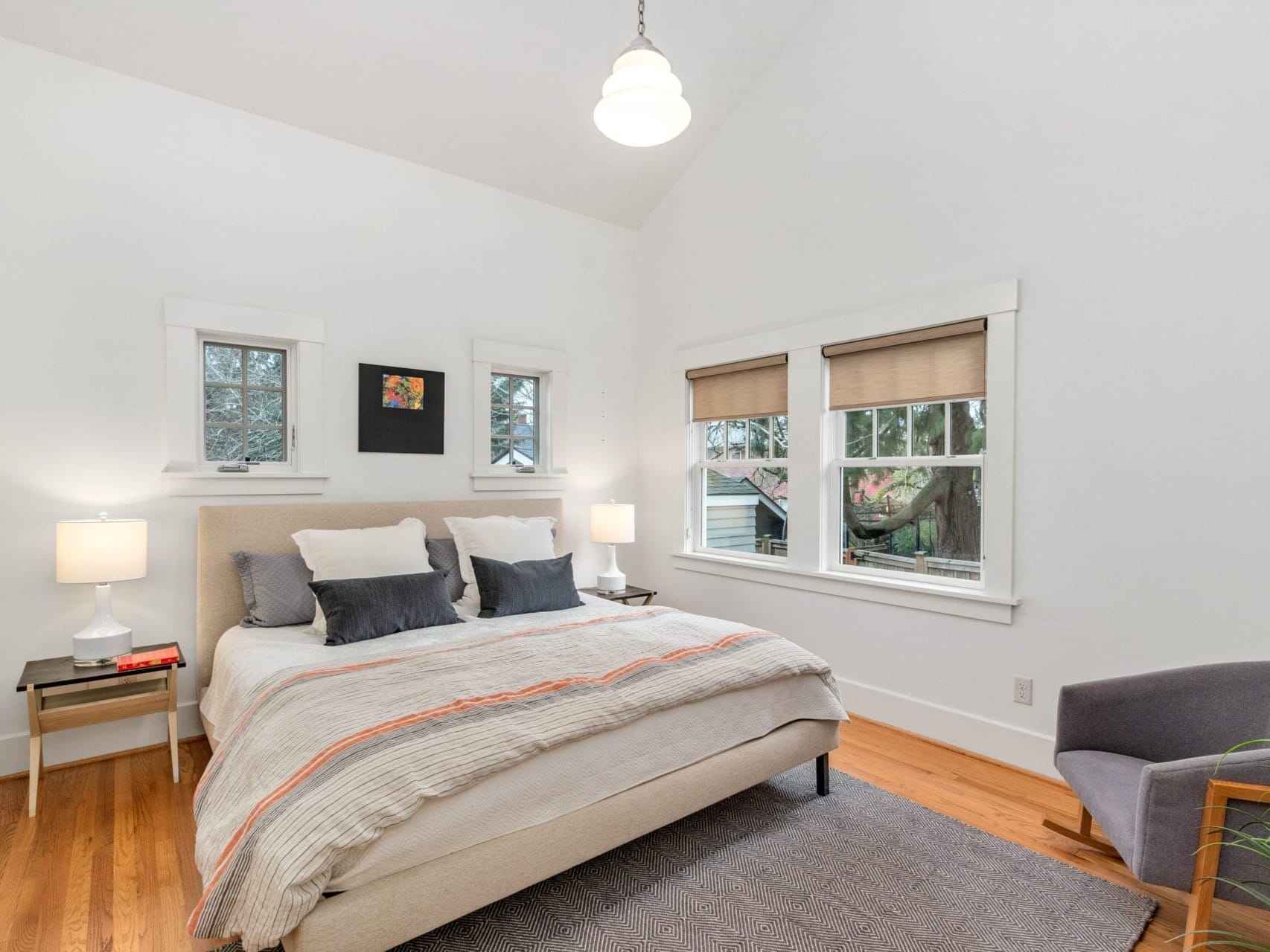 A cozy bedroom with a bed featuring gray and white bedding. Two small lamps are on nightstands beside the bed. A gray armchair is in the corner, and two windows with shade coverings offer natural light. A framed picture hangs above the bed.