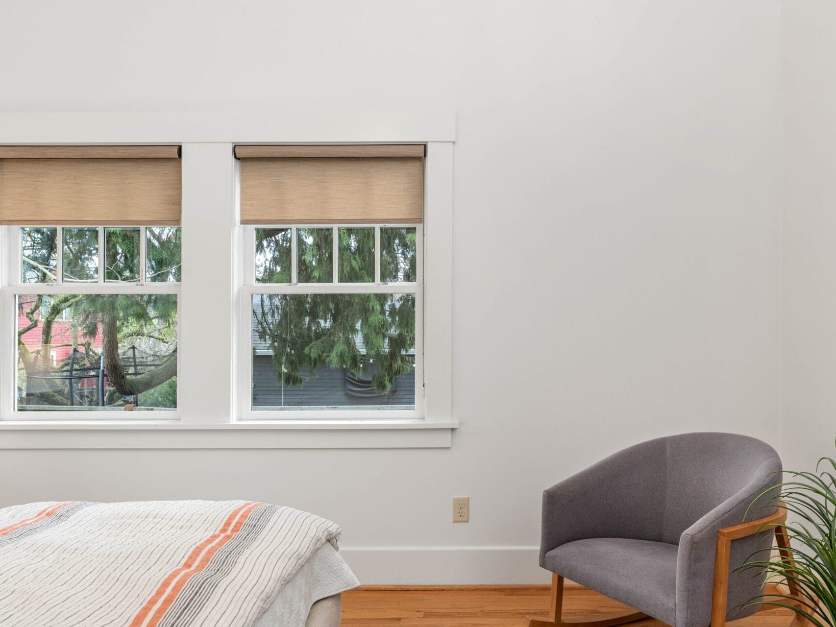 A minimalist bedroom with two large windows, each featuring beige roller blinds. A gray armchair sits in the corner on wooden flooring. A neatly made bed with a striped blanket is partially visible. Greenery can be seen outside through the windows.