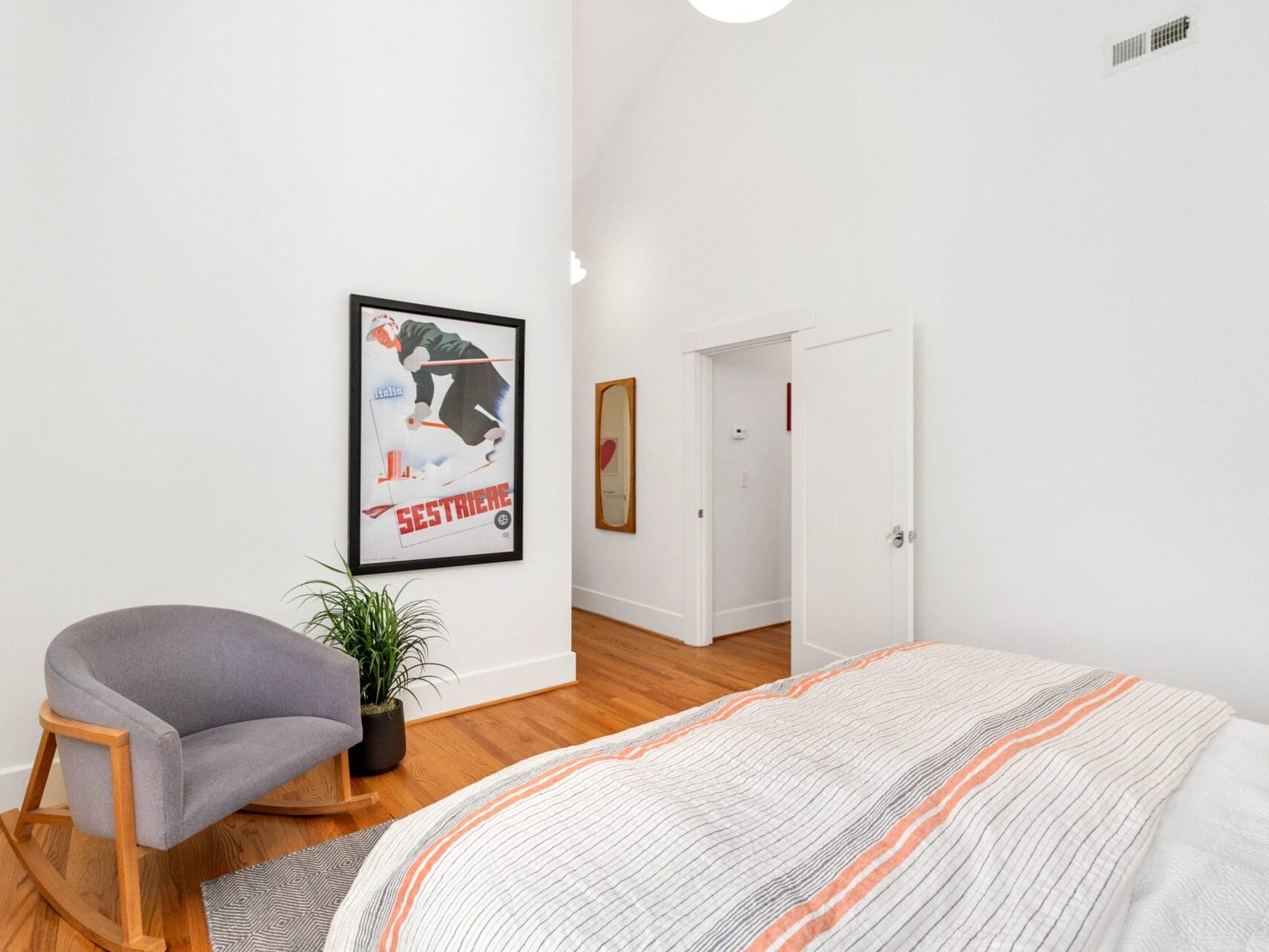 A minimalist bedroom with white walls features a framed artwork, a gray chair with a potted plant, and a bed with a striped comforter. Hardwood floors add warmth to the space. A partially open door is visible on the right.