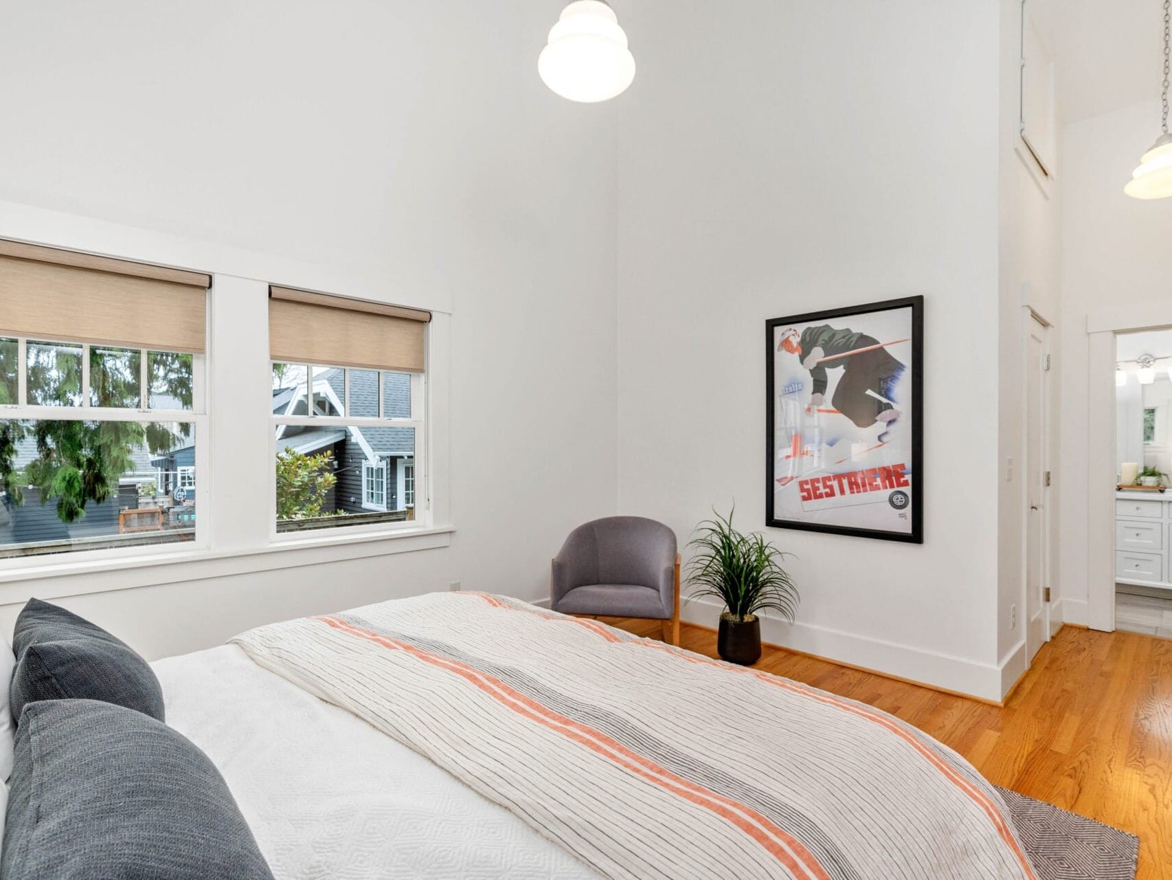 A bright bedroom with a large bed featuring white and orange bedding. Theres a gray armchair and a potted plant next to a framed poster on the wall. Two windows show a view of trees outside, and a glimpse into a bathroom is visible.