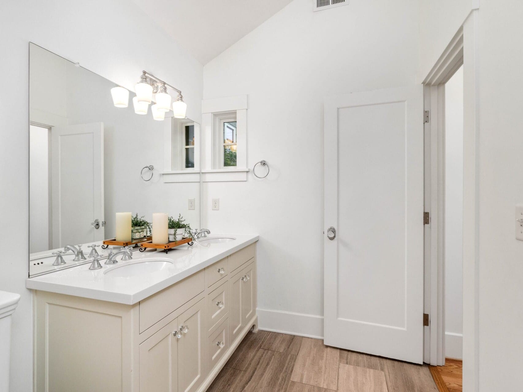 A bright, modern bathroom with a double sink vanity, large mirror, and overhead lights. The vanity has white drawers and candles on top. A closed door and towel ring are on the right wall. The floor is light wood.