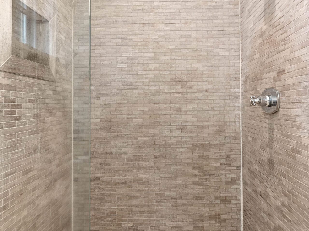 A minimalist shower with beige brick-style tiles on walls and floor, enclosed by a glass panel. It features a showerhead and a built-in shelf, creating a sleek, modern look.