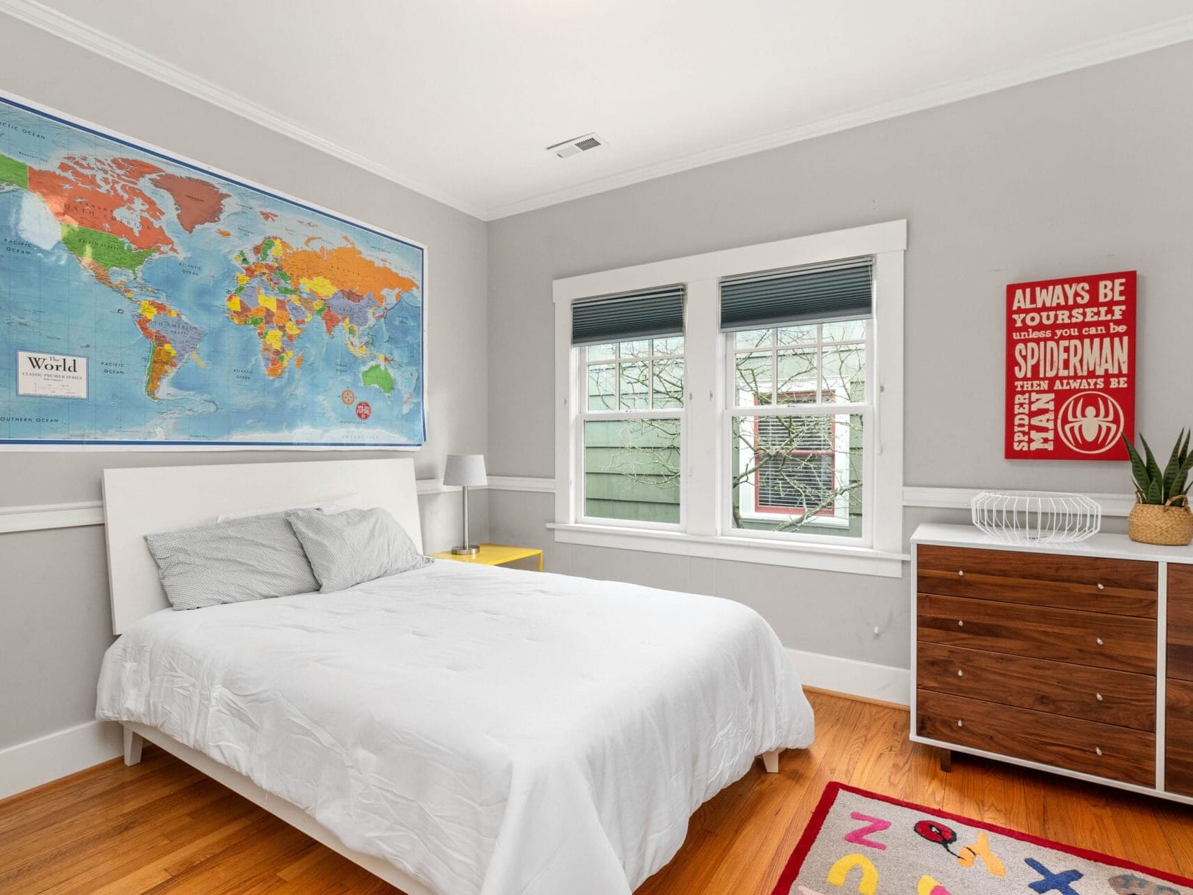 A bright bedroom with a white bed, light gray walls, and hardwood floors. A large world map and a poster are on the walls. A dresser with a plant is beside the bed, and a rug with colorful letters is on the floor. Two windows let in natural light.