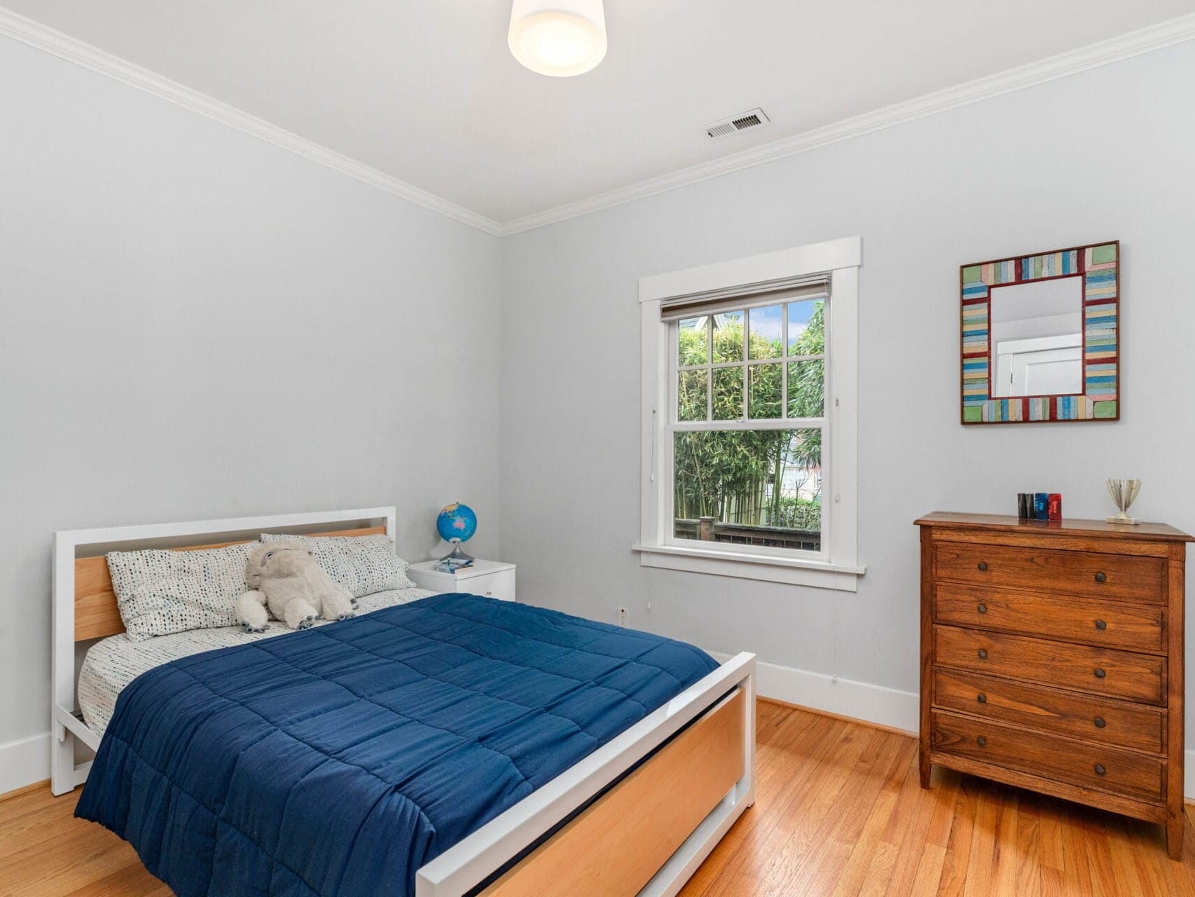 A simple bedroom with a wooden bed, blue bedding, and a teddy bear. A small globe sits on a white bedside table. A wooden dresser stands against the wall under a colorful framed mirror. A window shows greenery outside. The flooring is wooden.