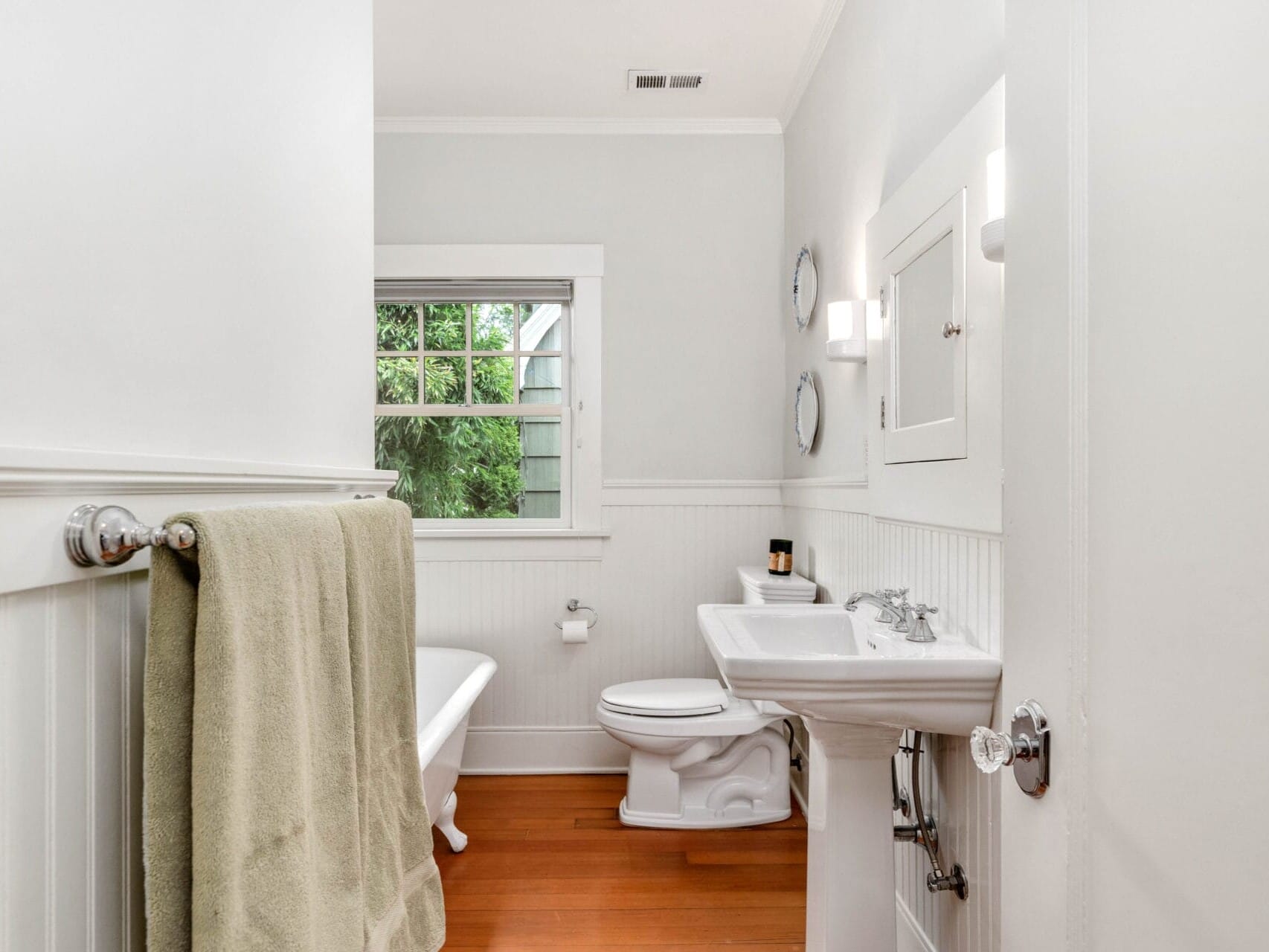 A bright bathroom features a white bathtub, a pedestal sink, and a toilet on a wooden floor. Above the sink are two circular mirrors and a small wall cabinet. A green towel hangs on a rail, and the window offers a view of greenery outside.