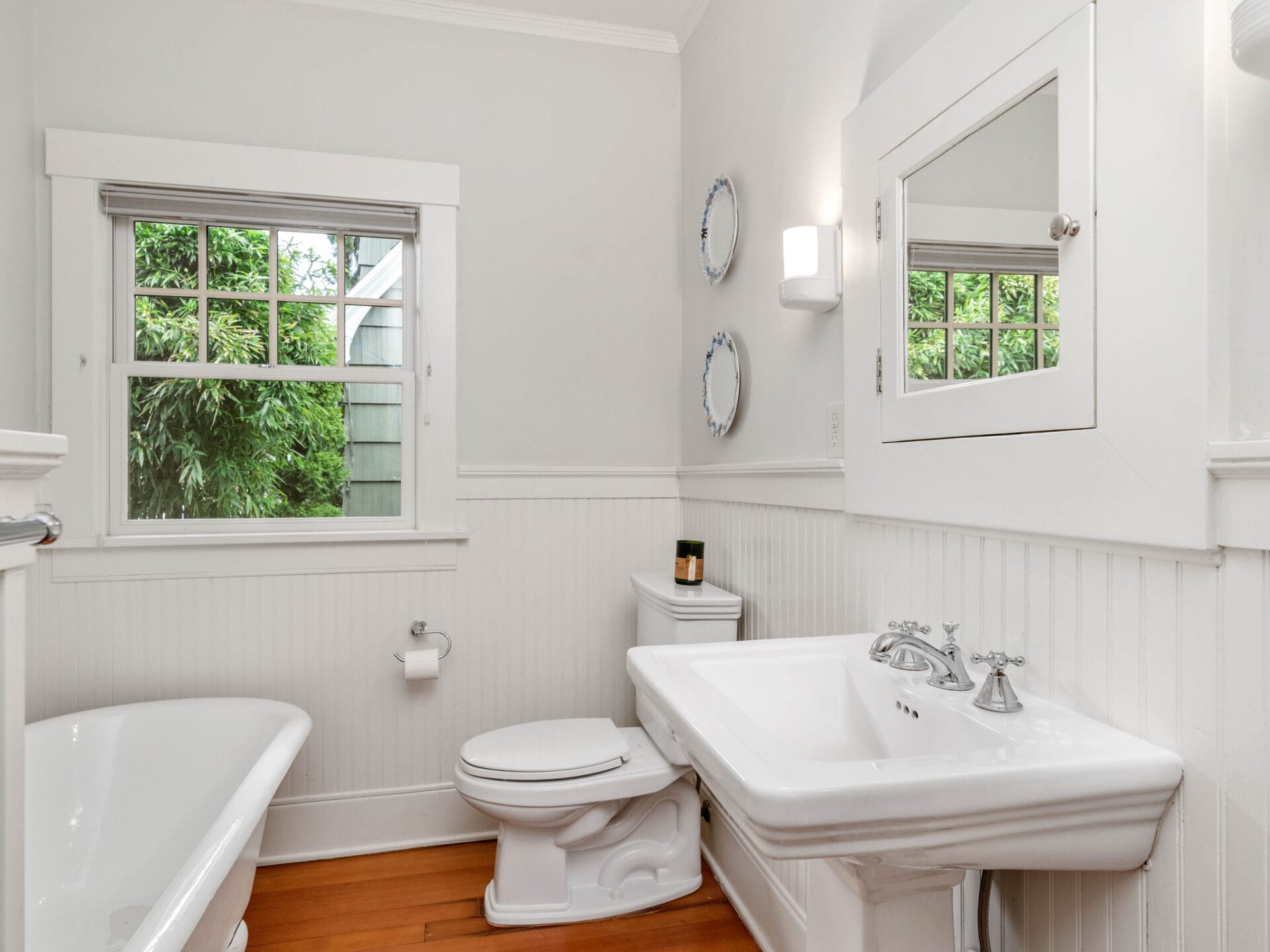 A bright, white bathroom with a window and natural light. Features a porcelain bathtub, toilet, and sink with chrome fixtures. Round mirrors and a candle adorn the space. White wainscoting decorates the walls, and light wood flooring complements the room.