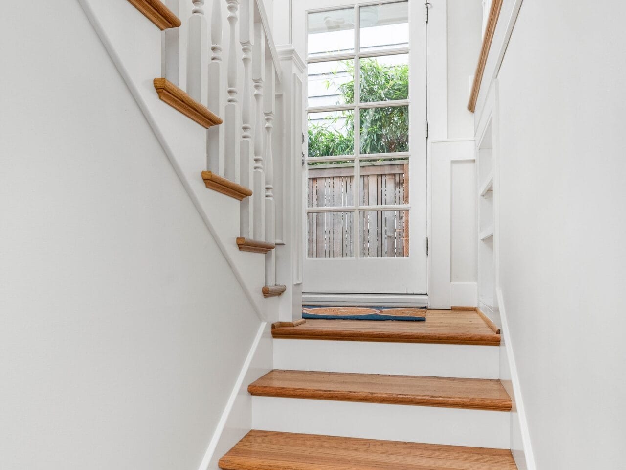 A staircase with wooden steps and white railings leads to a glass-paned door. A black-and-white framed photo hangs on the wall above the stairs. Natural light streams through the door, illuminating the space. A fence and greenery are visible outside.