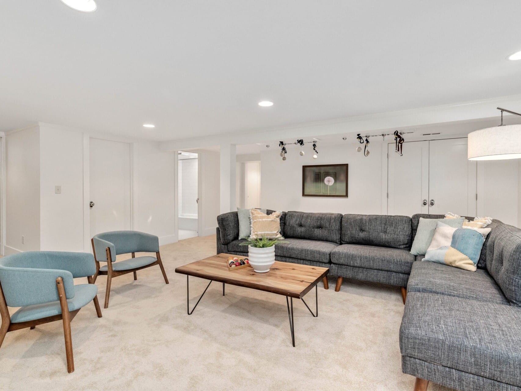 A modern living room with white walls and carpet, featuring a gray L-shaped sectional sofa. There are two blue upholstered chairs and a wooden coffee table. A potted plant, throw pillows, and wall art add decorative touches. Recessed and track lighting illuminate the space.