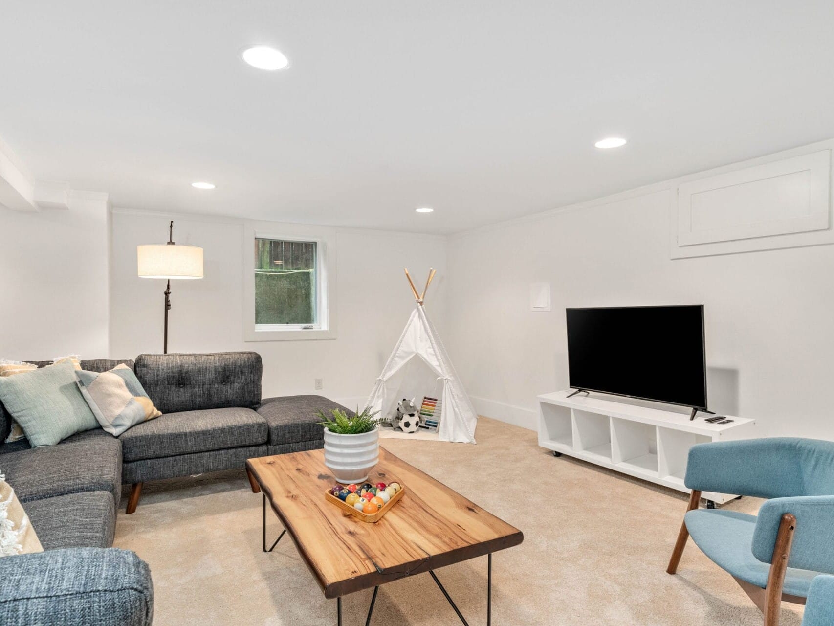 A cozy basement living room with a gray sectional sofa, a wooden coffee table, a large flat-screen TV on a white stand, and a small white play tent. The space is well-lit with recessed lighting, a floor lamp, and has a neutral decor.