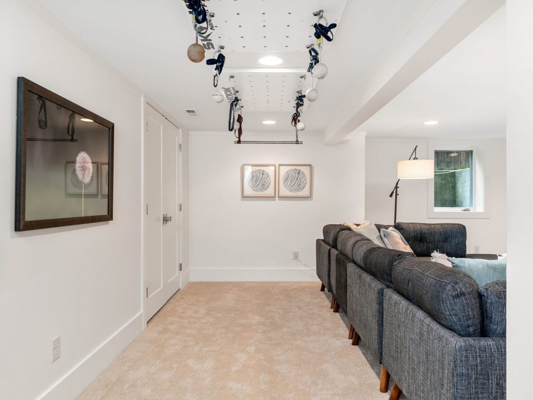 A cozy basement room with beige carpet and white walls. It features a dark gray sectional sofa, two abstract wall artworks, and unique ceiling decorations. The space is well-lit with a standing lamp and natural light from a small window.