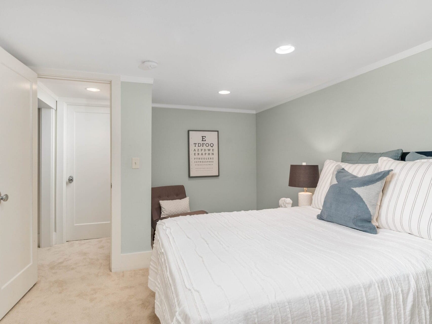 A bedroom with light gray walls features a neatly made bed with white linens and a gray throw pillow. A dark lamp is on the nightstand, and a brown chair sits against the wall near an inspirational poster. A door to the left is slightly ajar.