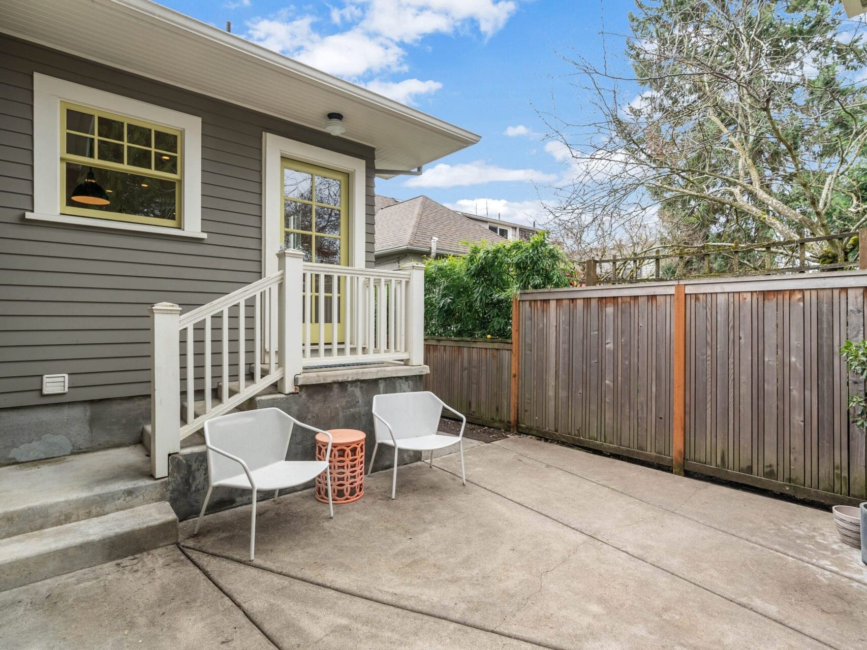 A small backyard patio with two white chairs and a small orange table on a concrete surface. A wooden fence surrounds the area, and a gray house with white trim is visible. Trees and a blue sky with clouds are in the background.