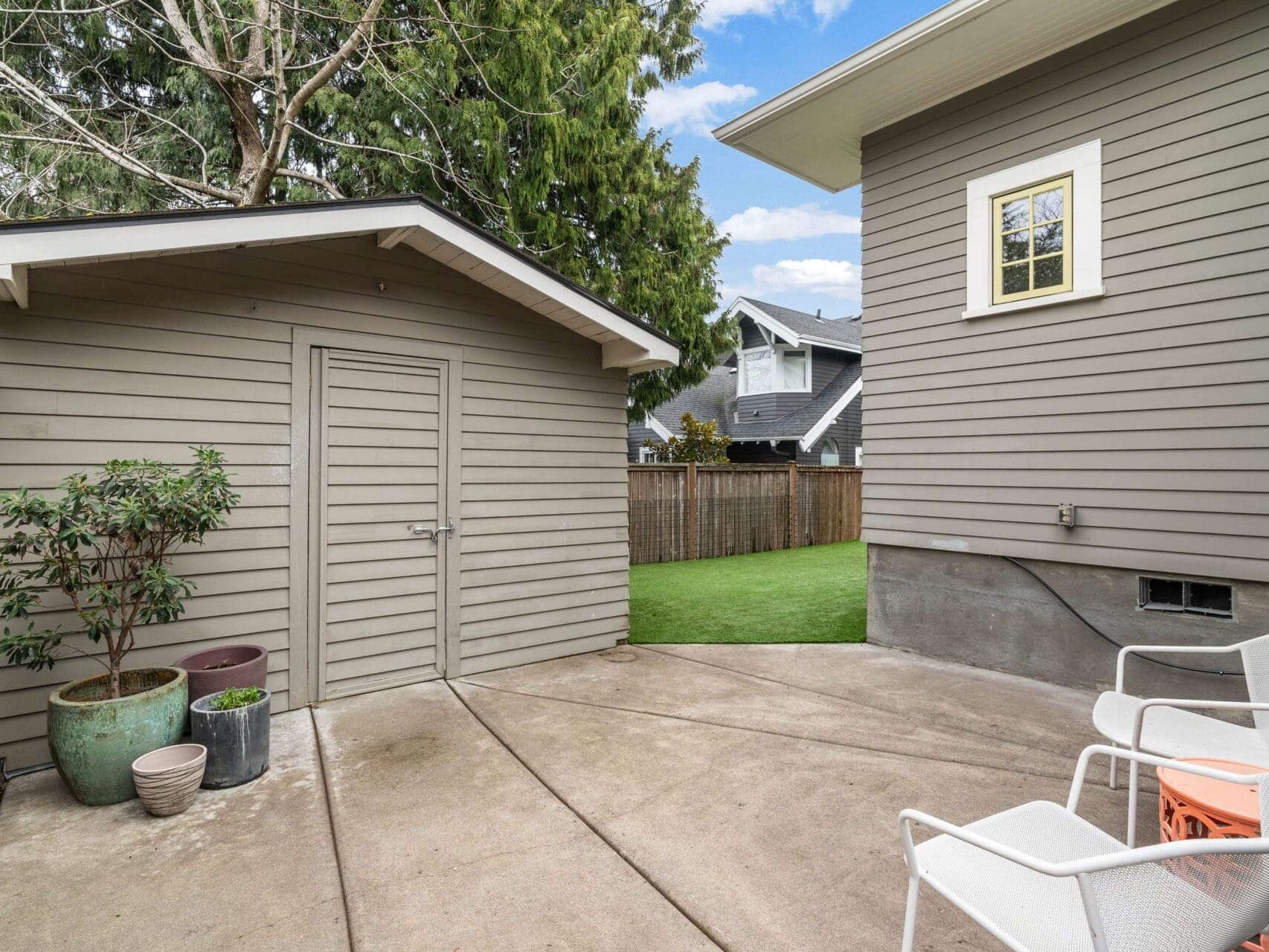 A backyard scene with a small shed next to a house. The area has concrete flooring with a few potted plants and several white chairs around a small table. A wooden fence and trees are visible in the background.