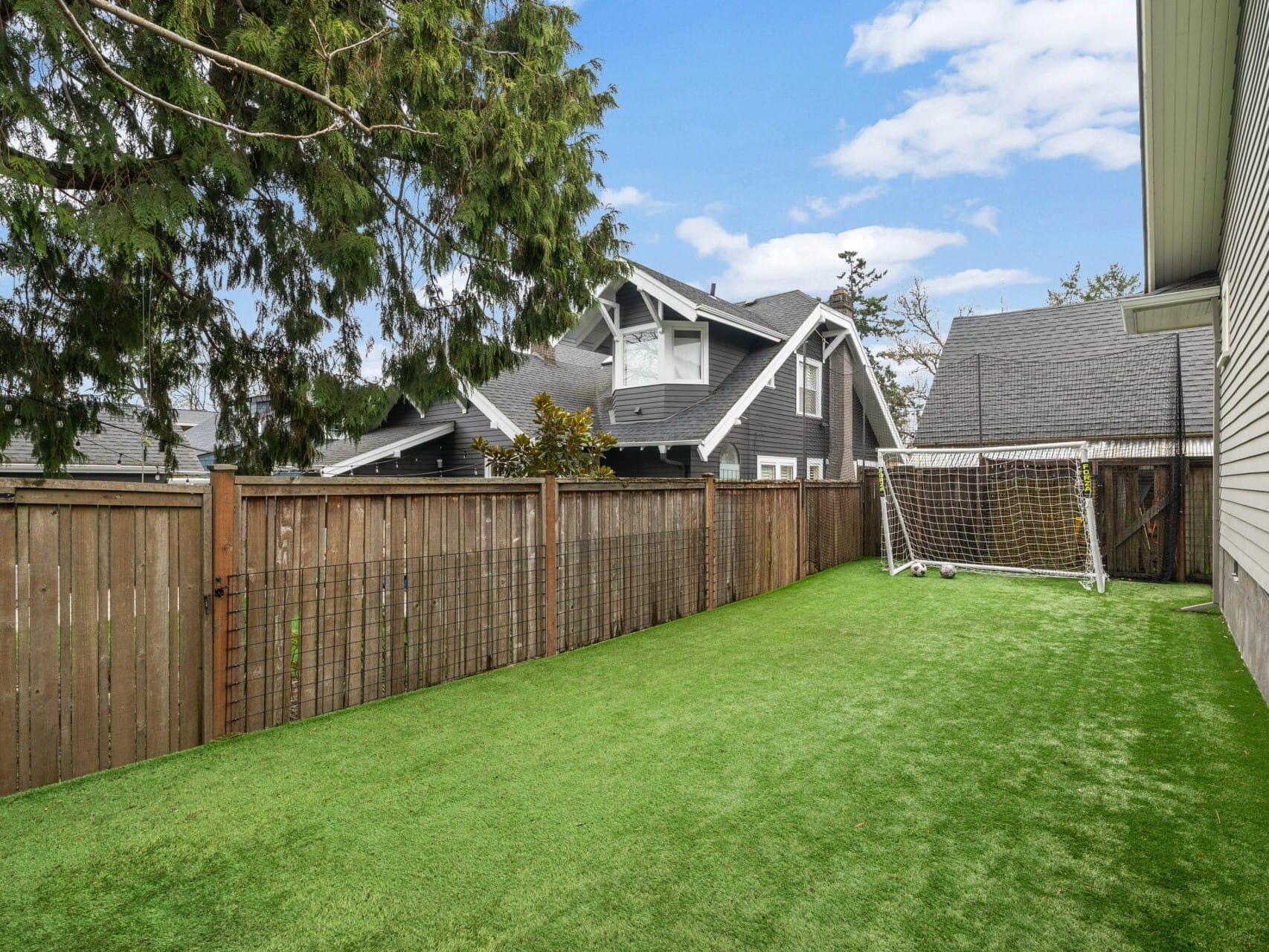 A backyard with artificial grass, a wooden fence, and a soccer goal. The area is bordered by two houses, and tall trees add greenery to the scene.